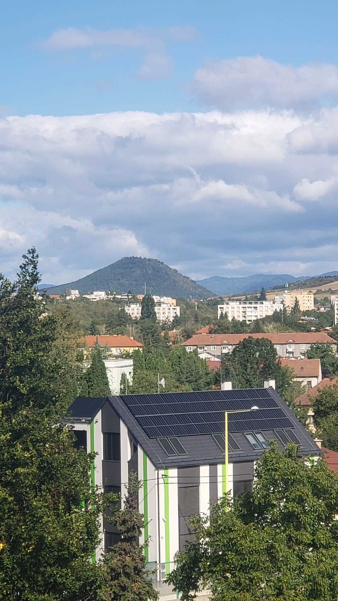 This is a vertical, medium-shot, eye-level outdoor photograph captured on a sunny day with scattered clouds. The foreground features a modern house with a dark, angled roof covered in solar panels and several skylights. The house has a distinctive design with vertical stripes of dark gray, white, and bright green. Trees with lush green foliage partially obscure the left and right sides of the house.
In the middle ground, a residential area with various buildings is visible, including houses with red-tiled roofs and several apartment buildings with white and light-colored facades. Some of these buildings are several stories high. The area is densely populated with trees interspersed among the structures.
The background is dominated by a large, tree-covered hill or mountain with a somewhat rounded peak. There appears to be a structure or small cluster of buildings on the summit of the hill. Beyond the hill, distant, rolling hills form the horizon under a bright blue sky with fluffy white and gray clouds.
This scene is likely located in Prešov, Slovakia, a city known for its hilly terrain and surrounding natural landscapes. The presence of residential buildings, apartment blocks, and the distinct geographical feature in the background are characteristic of the city.