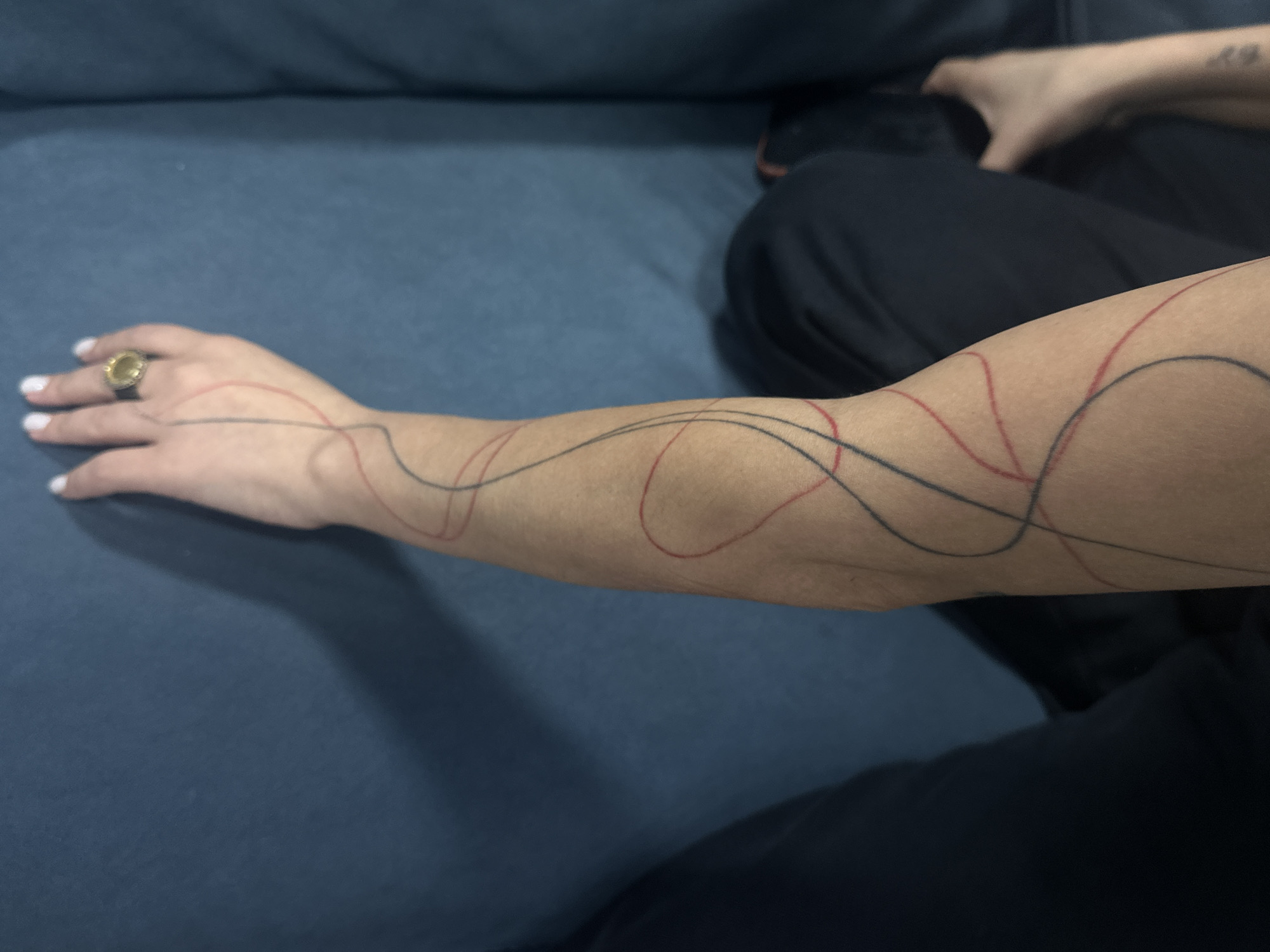 A person's arm is extended on a blue surface. The arm has a tattoo of wavy red and black lines. The person has a ring on their hand and is wearing black clothing. The location is unknown, and there is no evidence it is in Yerevan, Armenia.