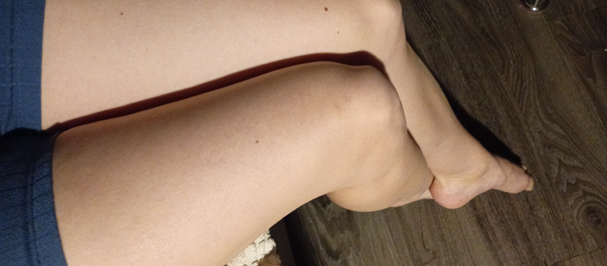 A photograph shows a person's legs. The legs are crossed at the knees. The background is a dark wooden floor. The person is wearing blue shorts. The image does not contain enough information to provide a location or other details.