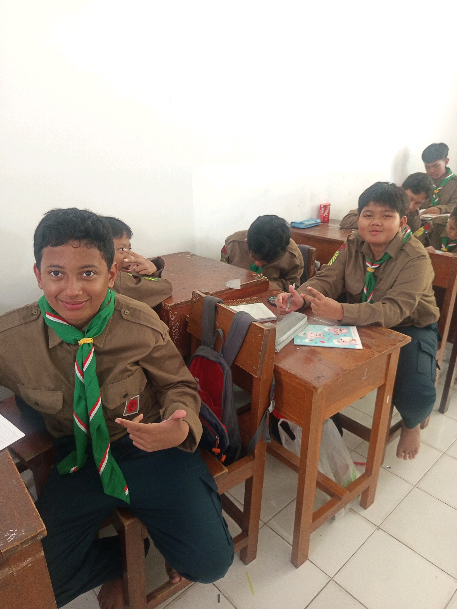 The image captures a classroom scene with young boys dressed in scout uniforms, likely in Malang, Indonesia, given the context. The boys are seated at wooden desks, some of which are arranged in rows. The focus is on two boys in the foreground. The boy on the left, facing the camera, is wearing a brown scout uniform with a green, red, and white scarf. He is smiling and gesturing with his right hand. Behind him and to his right, another boy in a similar uniform is looking towards the camera, holding a pen and making a peace sign with his fingers. Other boys are visible in the background, some engrossed in their studies, while others are partially obscured. The classroom environment appears simple, with white walls and tiled flooring. Objects like books, notebooks, and backpacks are present on the desks and floor. The overall atmosphere suggests a school day in session.