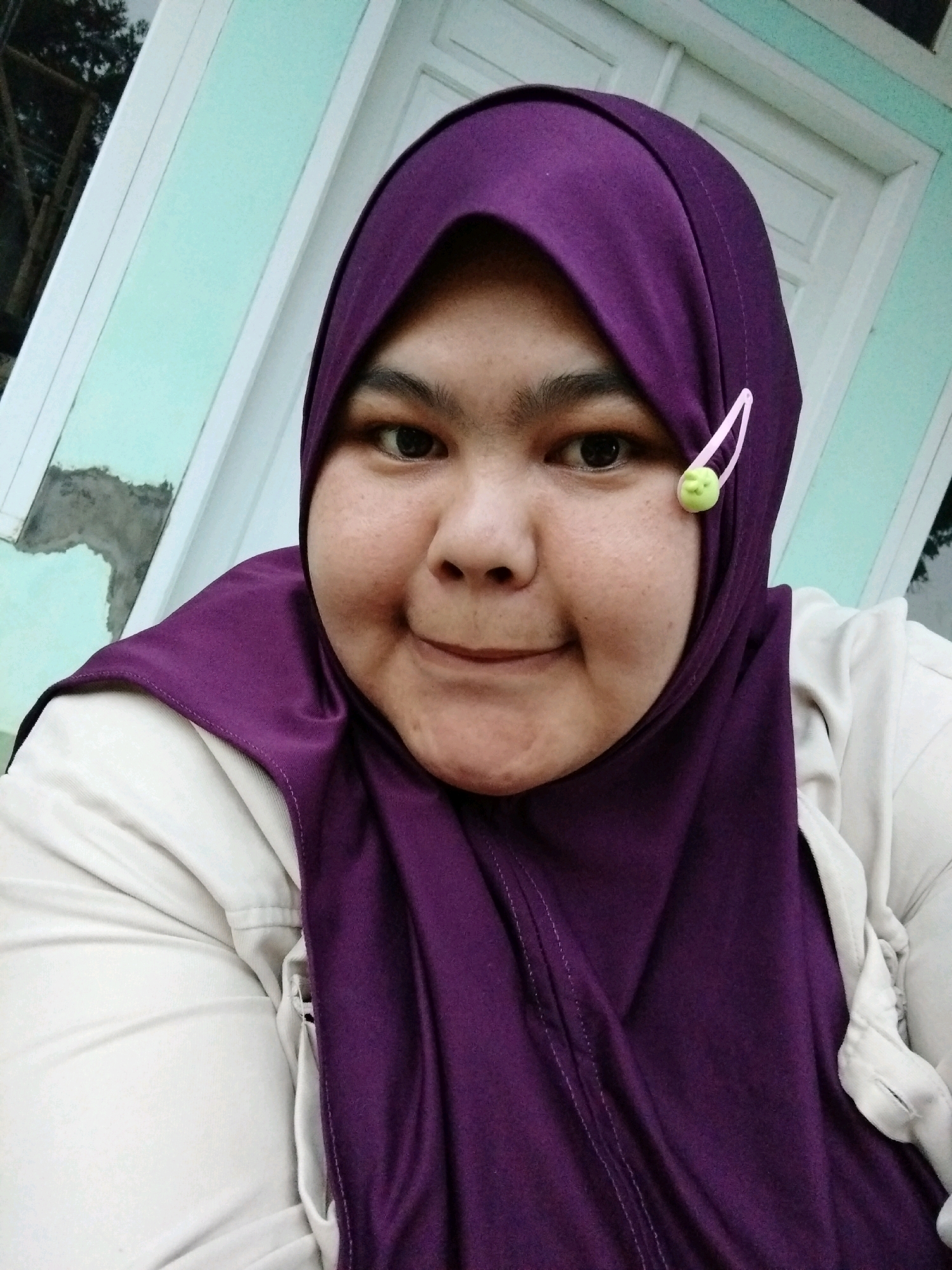 A young woman wearing a purple hijab and a light-colored jacket poses for a selfie in front of a wall with a turquoise-colored stripe and some visible mortar. A small green clip is attached to her hijab near her right eye. The background suggests an outdoor or semi-outdoor setting, possibly a home's exterior wall. The image appears to have been taken in Garut, Indonesia.