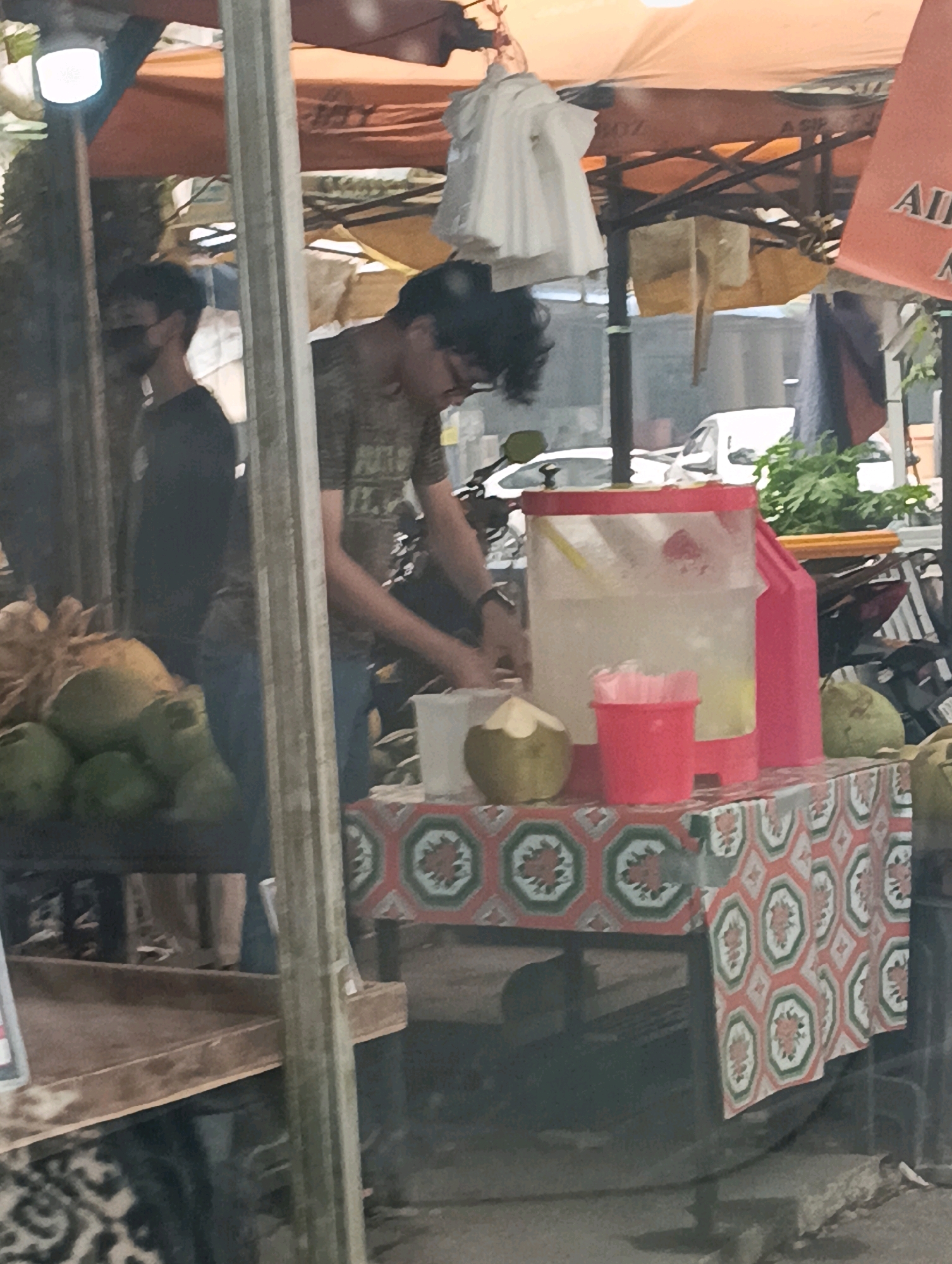 A medium-shot, eye-level, slightly blurry photograph captures a street scene in Kuching, Malaysia, beneath an orange awning. In the foreground, a young man with curly dark hair and glasses is partially visible, wearing a green and brown patterned t-shirt and blue jeans. He is standing behind a table adorned with a patterned cloth featuring red, green, and white octagonal designs with floral motifs. On the table, a large, clear plastic beverage dispenser filled with a light-colored liquid sits prominently. To the left of the dispenser is a small red cup filled with pink straws, and to the right, a large green pomelo. A half-cut green coconut is positioned in front of the dispenser, with another whole green coconut to its right. To the left of the frame, another young man, wearing a black t-shirt and a face mask, is visible, mostly obscured by a vertical metal pole. Behind the table, a white van is parked, and lush green foliage is visible in the background, hinting at an outdoor setting. The ground appears to be a mixture of concrete and dirt. The overall impression is of a roadside stall selling drinks and fruits.