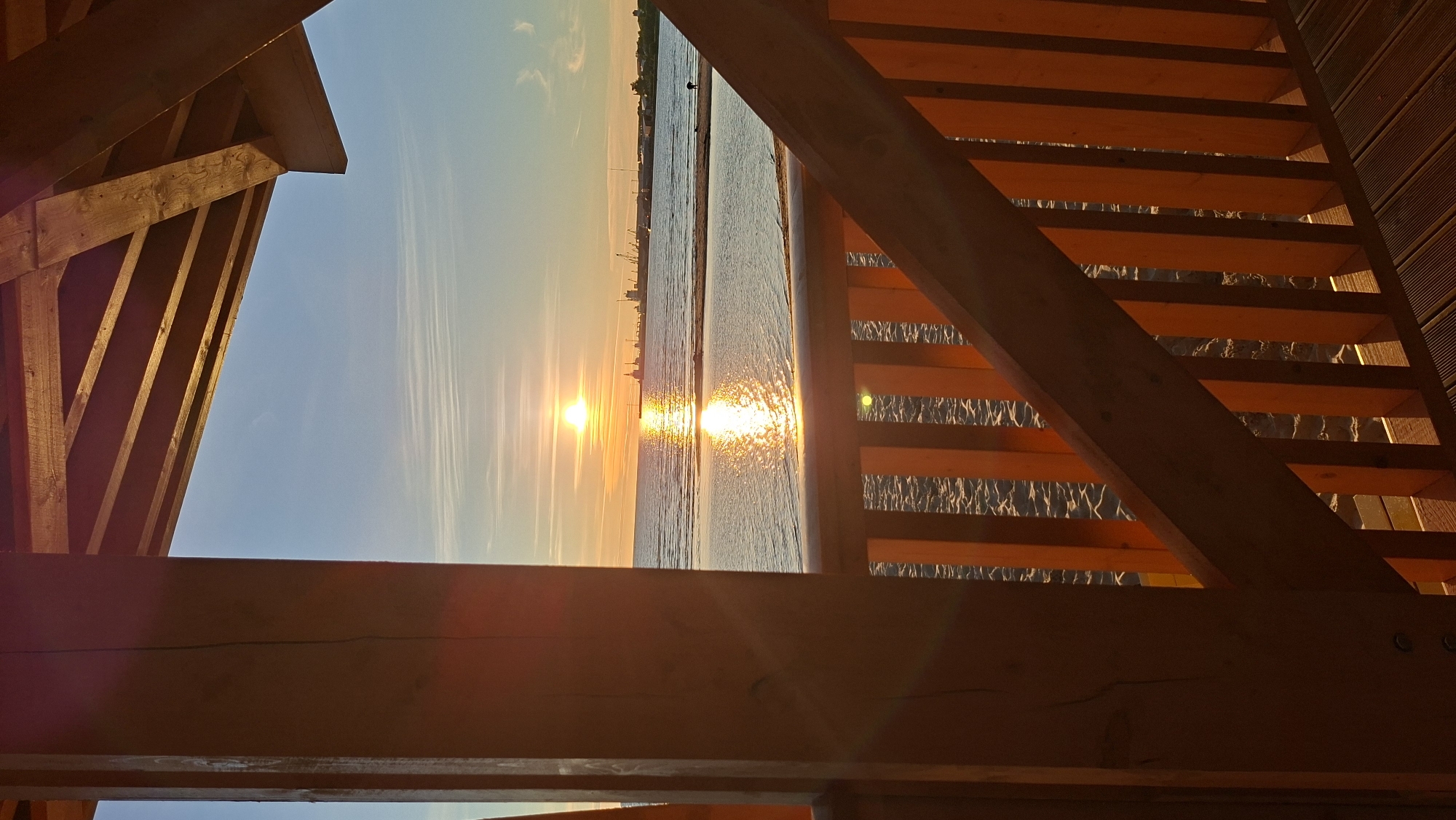 This is a vertical, medium shot photograph of a sunset over the Baltic Sea, viewed from within a wooden structure on the coast of Tallinn, Estonia. The sun, a bright orange orb, is low on the horizon, casting a brilliant reflection on the water. The sky is a gradient of pale orange and blue, with wispy white clouds. In the distance, a silhouetted industrial harbor with cranes is visible. The foreground is dominated by the warm-toned wooden structure, with thick vertical beams and horizontal planks forming a railing and part of a roof. The beach, with scattered sand and pebbles, is visible below the railing. A single, dark figure, possibly a person, stands on the beach in the mid-ground, facing the sea. The lighting suggests it is late afternoon or early evening, with the golden hour casting a warm glow on the scene.
