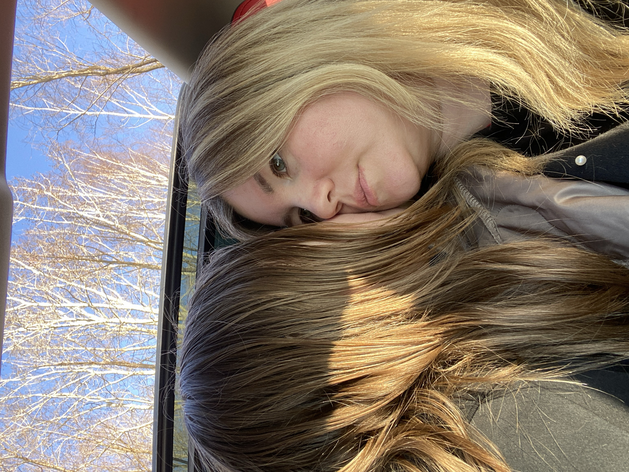 An image captured from a slightly overhead perspective shows two individuals, likely women, inside what appears to be a vehicle, possibly a car or bus. The person in the foreground has long, wavy blonde hair with lighter highlights, a fair complexion, and green or hazel eyes, looking gently towards the left. Her head is resting on the abundant, slightly darker brown hair of a second, mostly unseen individual beneath her. Her expression appears calm and relaxed.
She is wearing a dark top or jacket, with a small white, pearl-like button visible on the collar area. A hint of a red object, possibly a headband or headphones, can be seen partially obscured by her blonde hair near the top of her head.
Through a window on the left side of the frame, a clear, bright blue sky is visible, along with numerous bare trees, their branches creating an intricate pattern against the sky. This suggests a season of late autumn, winter, or early spring. Strong sunlight illuminates the scene, casting bright highlights on the hair of both individuals, indicating it is daytime, likely midday or early afternoon. The location context for this image is Chaikovsky, Russia. No visible text is present in the image.