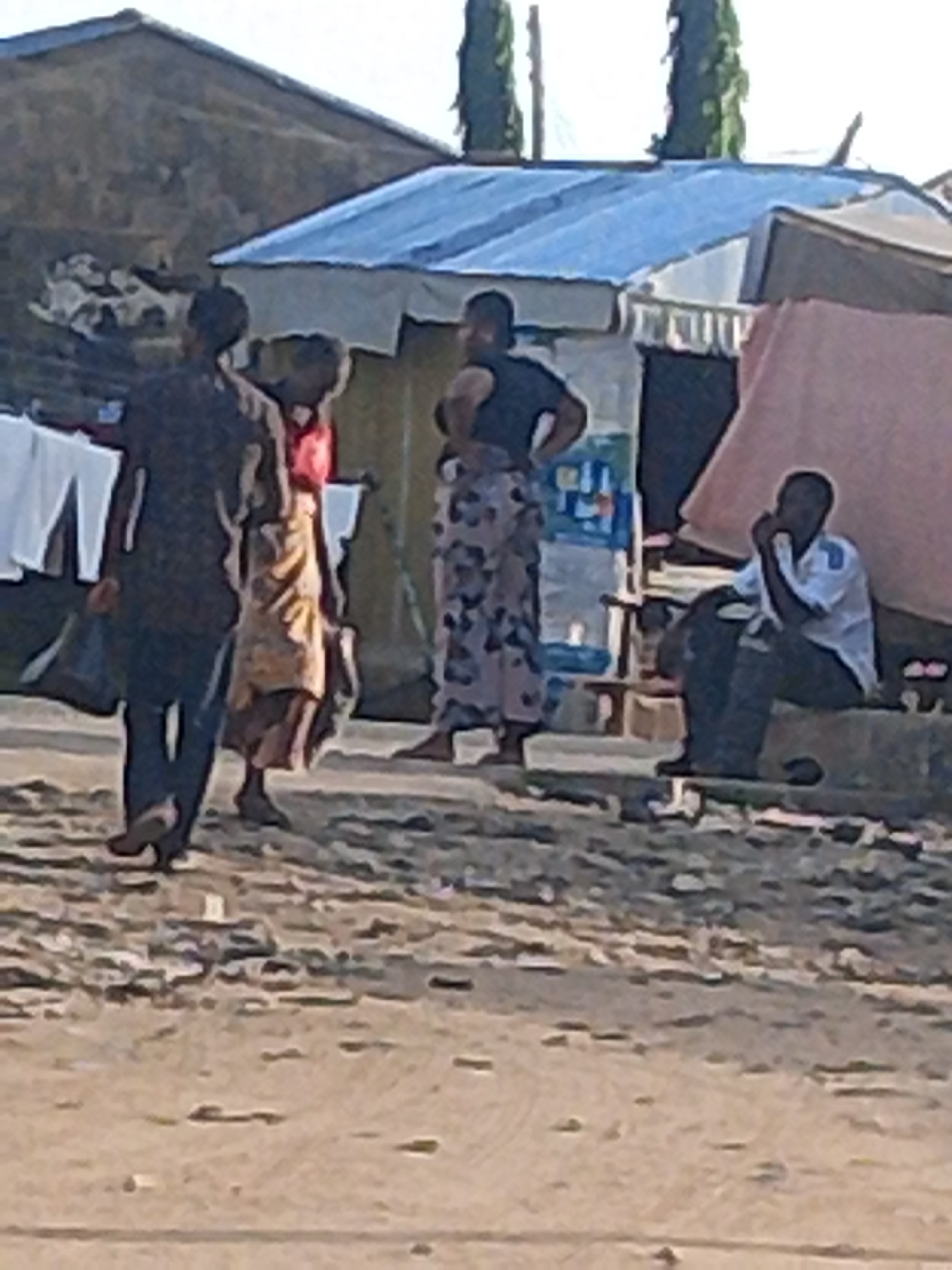The image depicts a street scene in Jalingo, Nigeria, likely during the daytime given the lighting. Four individuals are visible. In the foreground, two people are walking from left to right. The person on the left wears a dark top and dark pants, carrying a dark bag. The person next to them, slightly behind, wears a brightly colored patterned skirt and a reddish top.
Further back, a person in a dark top and a patterned skirt stands facing left, with their hands on their hips. To the right, a person is seated on a low concrete structure. This seated individual wears a light-colored shirt and dark pants and appears to be holding something to their mouth or chin.
Behind the people, several structures with corrugated metal roofs are visible, suggesting a built-up area that may be a market or a residential zone. Laundry is hanging on a line in the background to the left. The ground appears to be dirt or gravel. The overall atmosphere suggests a casual, everyday moment. No visible text is present in the image.