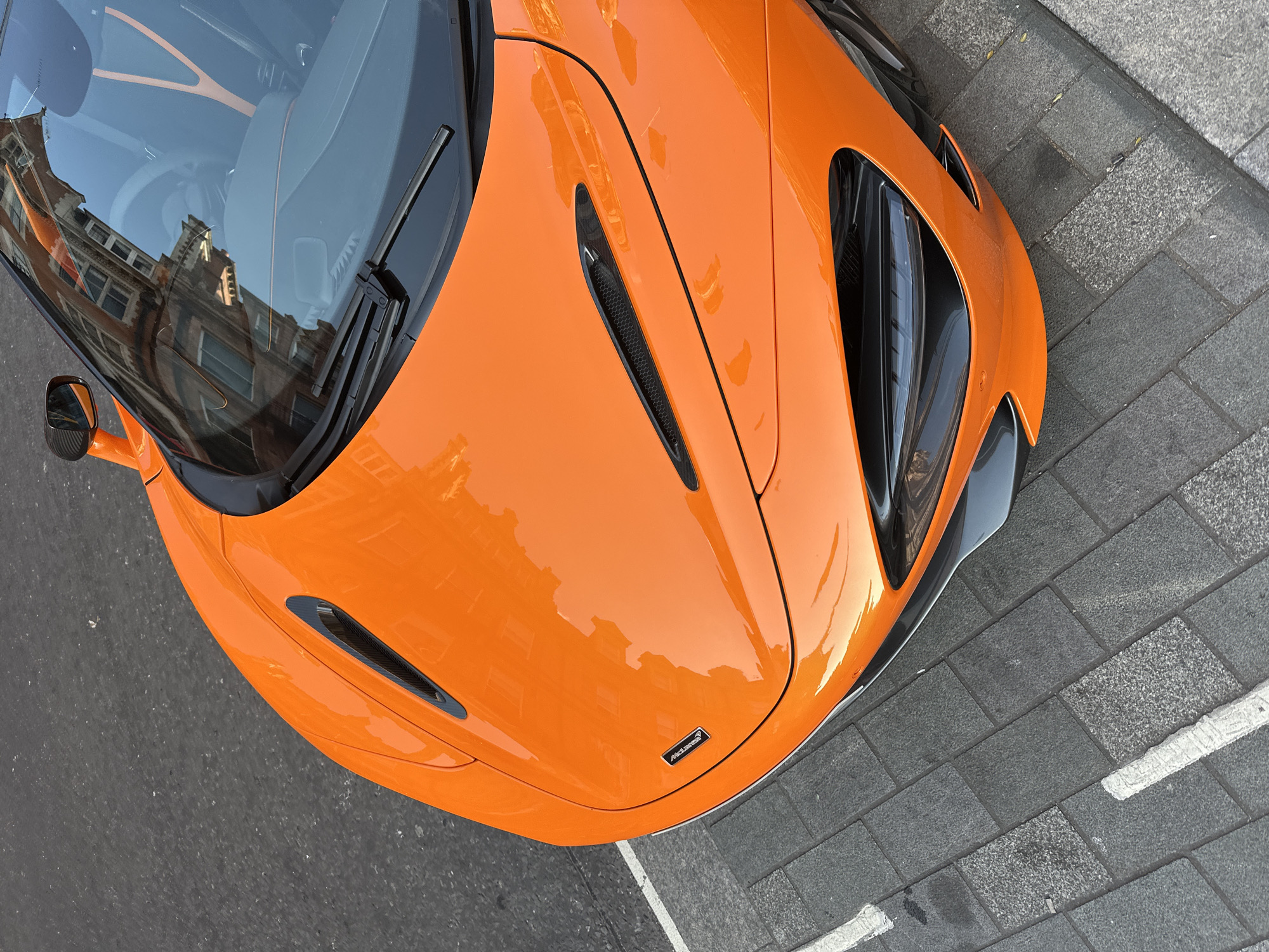 The image displays an overhead, slightly angled view of the front section of a vibrant orange McLaren supercar, parked on an urban street. The car's highly reflective, glossy finish prominently mirrors its surroundings. The large windshield captures a clear reflection of a multi-story building constructed of reddish-brown brick with numerous windows, indicative of classic city architecture often found in London. A bright sky is also reflected, suggesting clear daylight conditions. Faint details of the car's interior, including a light-colored steering wheel and orange dashboard elements, are visible beneath the reflections. Black windshield wipers are positioned at the base of the screen.
The car's aggressive design incorporates several black, honeycomb-patterned grilles and vents, including a prominent elongated vent on the hood and larger air intakes integrated into the dark grey lower front bumper and side panels, emphasizing its high-performance nature. A subtle, black "McLaren" badge is visible on the center of the front hood.
The vehicle is situated with its right side adjacent to a grey block-paved sidewalk, marked with a partial white line, while its left side extends over a darker asphalt road surface. No individuals are present in the scene. The overall impression is that of a powerful, luxurious sports car resting in a European city environment under bright, daytime conditions.
