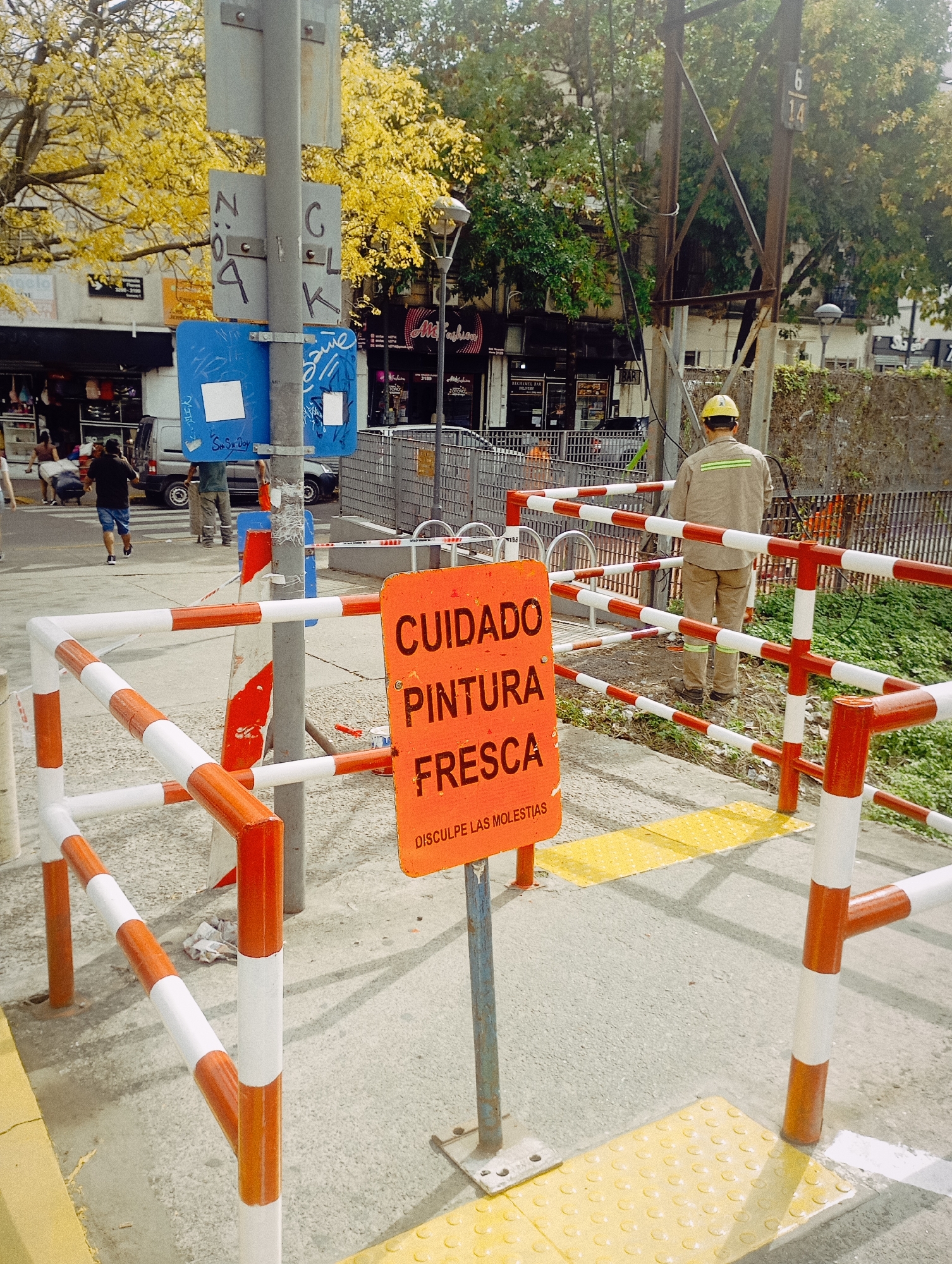 The image shows a street scene in Buenos Aires, Argentina. In the foreground, there is a sign in Spanish that reads "Cuidado Pintura Fresca" (Caution Fresh Paint) and "Disculpe las Molestias" (Sorry for the Inconvenience). The sign is mounted on a metal pole and surrounded by a barrier with orange and white striped bars. To the right of the sign, a worker wearing a yellow hard hat and beige work clothes is standing on a yellow tactile paving surface. Behind the barrier, there is a metal fence and some greenery. In the background, a street with shops and people can be seen. There is a utility pole with a number 6/14 and various signs attached to a pole, including the word "NO". Cars are parked along the street, and pedestrians are walking. The overall setting is an urban environment with trees and buildings.