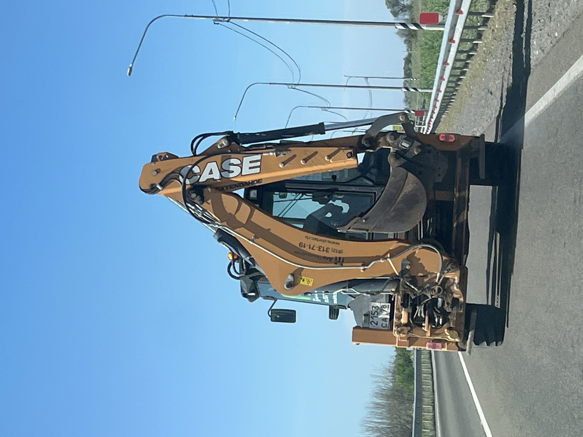 An orange backhoe loader with the brand name "CASE" visible on the boom is driving on a paved road under a clear blue sky. The backhoe is facing away from the camera, with its boom extended upwards and forward. The license plate of the vehicle reads "2153 CA 78". On the side of the boom, there is a sticker with a phone number "(812) 313-71-19" and a website "www.dortec.ru". To the right of the road, there is a metal guardrail with black and white striped poles. In the distance, there are trees and green fields. The scene is consistent with a road in Bakhtemir, Russia.
