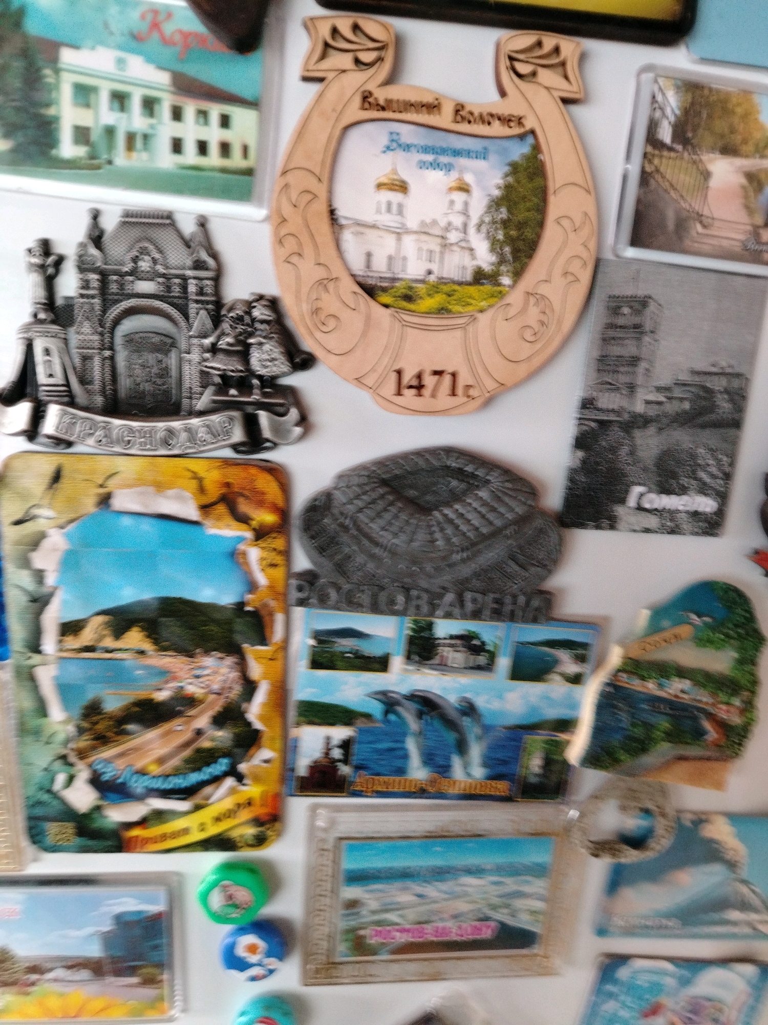 The image shows a collection of refrigerator magnets on a white surface, likely a refrigerator door. The magnets depict various landmarks and scenes.
One magnet features a wooden horseshoe-shaped frame with an image of a white church with golden domes. The text on the magnet reads "Вышний Волочек" (Vyshny Volochek) and "Воскресенский собор" (Resurrection Cathedral), and "1471". This indicates the magnet is a souvenir from Vyshny Volochek, Russia, and refers to the Resurrection Cathedral, with a date of 1471.
Another magnet is a silver-toned metal relief of an arched structure with two figures in front of it, and the word "КРАСНОДАР" (Krasnodar) inscribed below. This magnet is a souvenir from Krasnodar, Russia.
There are also several photographic magnets. One shows a coastal scene with a road and a beach filled with people. Another depicts jumping dolphins. There are also magnets with images of buildings and landscapes.
The image does not contain any individuals or specific actions. The environment is a plain white background, suggesting it's a close-up shot of a refrigerator. There is no visual information present that specifically relates to the city of Pregradnoye, Russia.