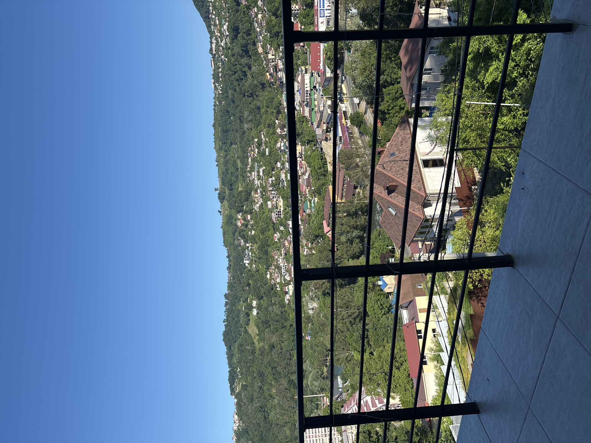 The image shows a view from a balcony overlooking a hillside town in Sochi, Russia. The foreground features a tiled balcony floor and a black metal railing. Beyond the railing, a dense green hillside is covered with numerous buildings, ranging from single-family homes to larger apartment complexes. Many of the buildings have red-tiled roofs. Trees are interspersed throughout the hillside, creating a lush, green landscape. The sky is a clear, bright blue. In the middle ground, there appears to be a road with some commercial buildings and a river or body of water winding through the town. The overall scene depicts a vibrant, inhabited hillside community under a sunny sky.