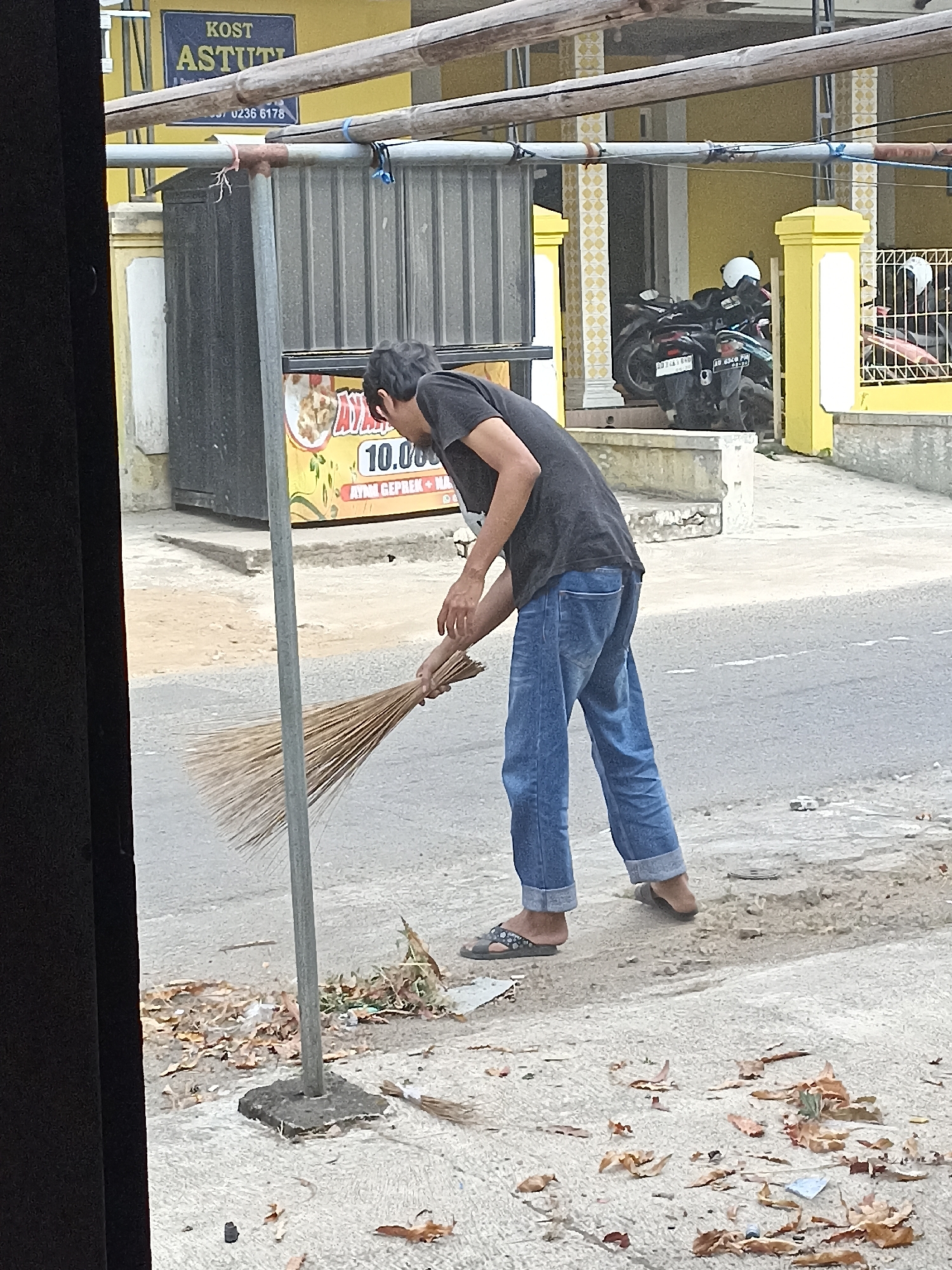 In the city of Pulisen, Indonesia, a man wearing a dark t-shirt and blue jeans is sweeping the ground with a broom made of straw. He is standing on a paved surface next to a street, with fallen leaves scattered around his feet. In the background, there is a building with a yellow facade, a metal fence, and several motorcycles parked. A sign with text "KOST ASTUTI" is visible above the building. The scene appears to be a street or sidewalk area, possibly in a residential or commercial district.