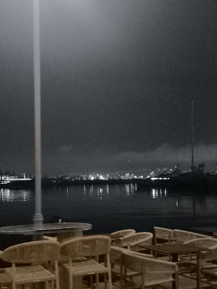 This nighttime photograph presents a tranquil waterfront scene in Palaio Faliro, Greece. The foreground features an outdoor seating area, equipped with several empty rattan-style chairs and tables, suggesting a quiet moment at a cafe or restaurant. A tall, light-colored pole stands prominently on the left side, extending vertically through the frame.
Beyond the seating area, a calm body of water stretches across the midground, serving as a reflective surface for the distant city lights. Across the water, the background reveals a cityscape with numerous twinkling lights, indicating a populated area nestled against a hill or shoreline. On the right side of the water, the tall mast of a sailboat is visible, silhouetted against the dark sky. On the left, a pier or dock area with some illumination can be seen.
The sky is dark and overcast with visible clouds, contributing to the atmospheric nighttime setting. No people or specific activities are depicted, giving the scene a serene and almost contemplative mood. The image has a desaturated aesthetic, dominated by dark blues and grays, with the chairs and tables offering a warm, muted brown tone. No visible text is present.