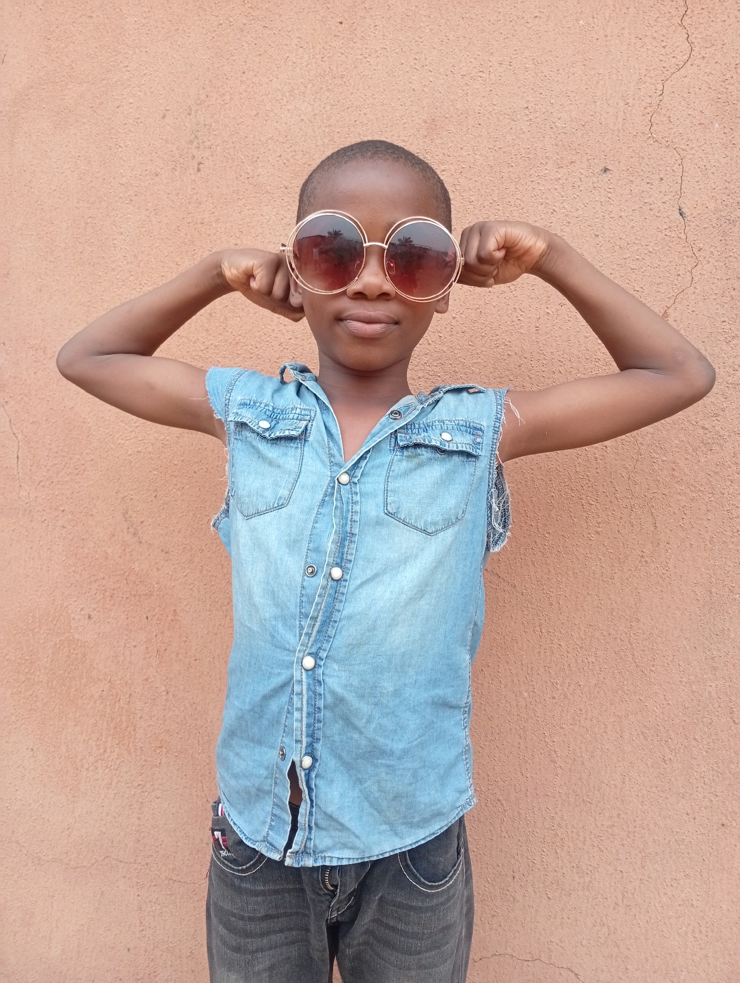 A young, dark-skinned child stands centered in front of a mottled reddish-brown wall. The child is wearing large, round sunglasses with gold frames and dark lenses. The child is also wearing a light blue denim vest over dark jeans. The vest is buttoned but the last button is undone. The child has both arms raised, with bent elbows, hands near their face appearing to hold the sunglasses in place. The background wall has minor cracks and texture. The scene appears to be outdoors in daylight. The location is Sabon Tasha, Nigeria.