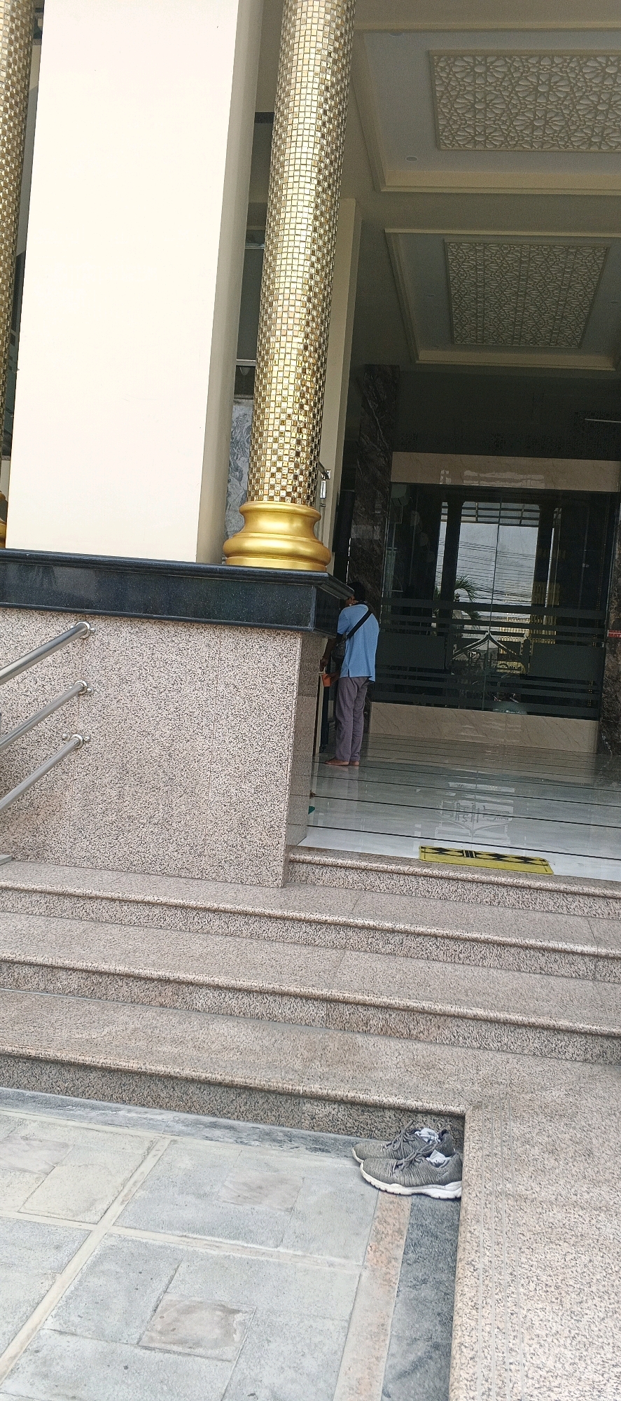 In the city of Sumberwudi, Indonesia, a man wearing a light blue t-shirt and gray pants stands facing a glass door entrance to a building. He is positioned on a paved surface at the bottom of a granite staircase, with his back mostly to the camera. A pair of gray athletic shoes with white laces and some crumpled white paper inside are placed on the ground to the right of the stairs. The building entrance features large glass doors framed by dark marble. In front of the entrance, a yellow doormat is visible. To the left of the man, a large, cream-colored column is adorned with gold mosaic tiles and has a golden base. The surrounding environment consists of granite-clad walls and marble accents on the building's exterior. The scene appears to be outdoors, with the time of day suggested by the lighting to be daytime.