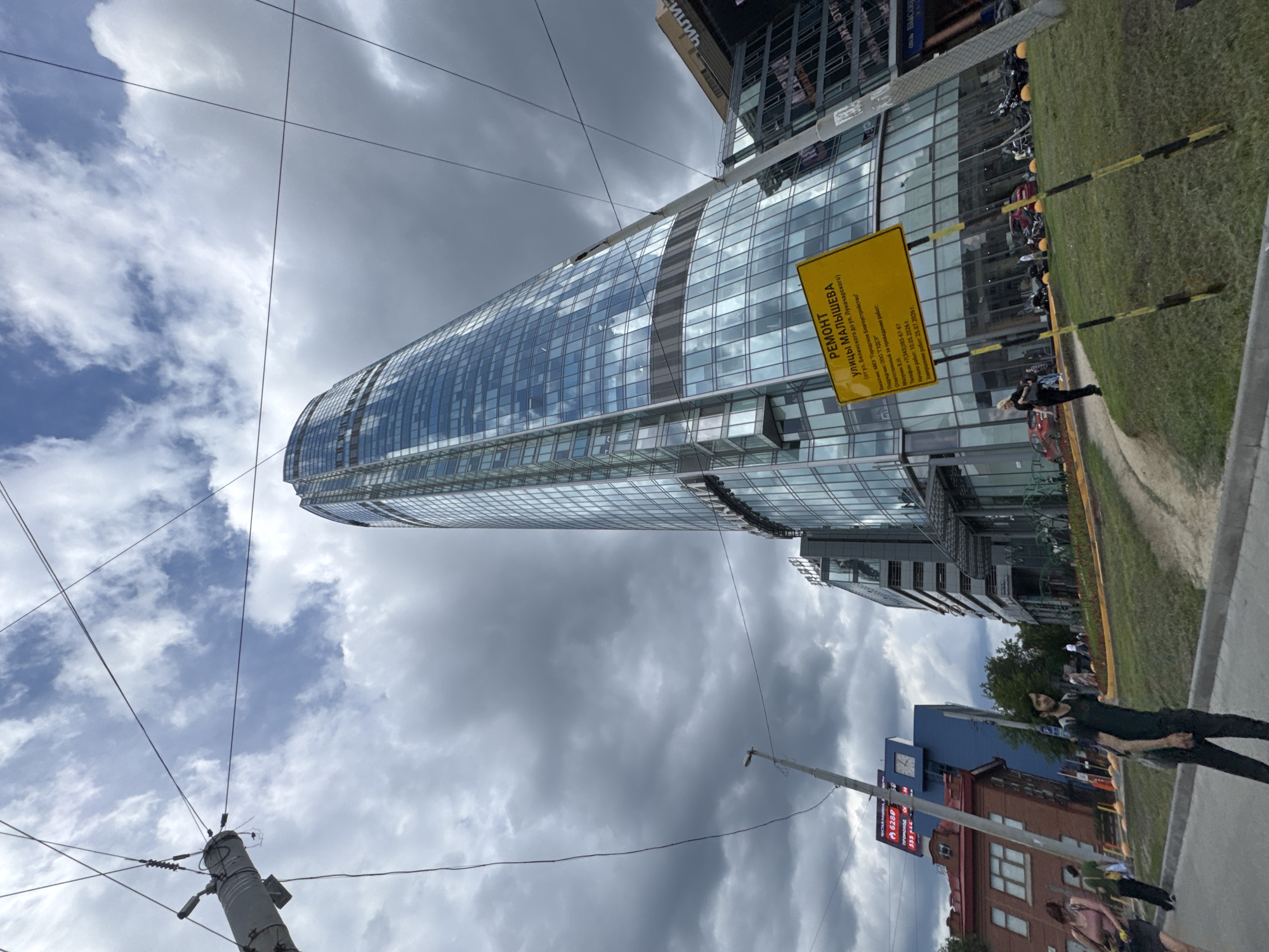 An exterior, low-angle, wide shot captures a modern, glass-clad skyscraper in Yekaterinburg, Russia, under a cloudy sky. The building, which appears to be the Vysotsky Business Center, dominates the right side of the frame. In the foreground, a dirt path curves through a grassy area. Several utility poles and wires crisscross the sky, obscuring portions of the building and clouds. A yellow sign with Cyrillic text, indicating road repairs on Malysheva Street, is prominently displayed on the right. In the mid-ground, several people are visible walking on a sidewalk and near parked motorcycles. To the left of the skyscraper, a smaller red brick building and a blue building with a clock face are visible. The overall scene depicts an urban environment with ongoing infrastructure work.