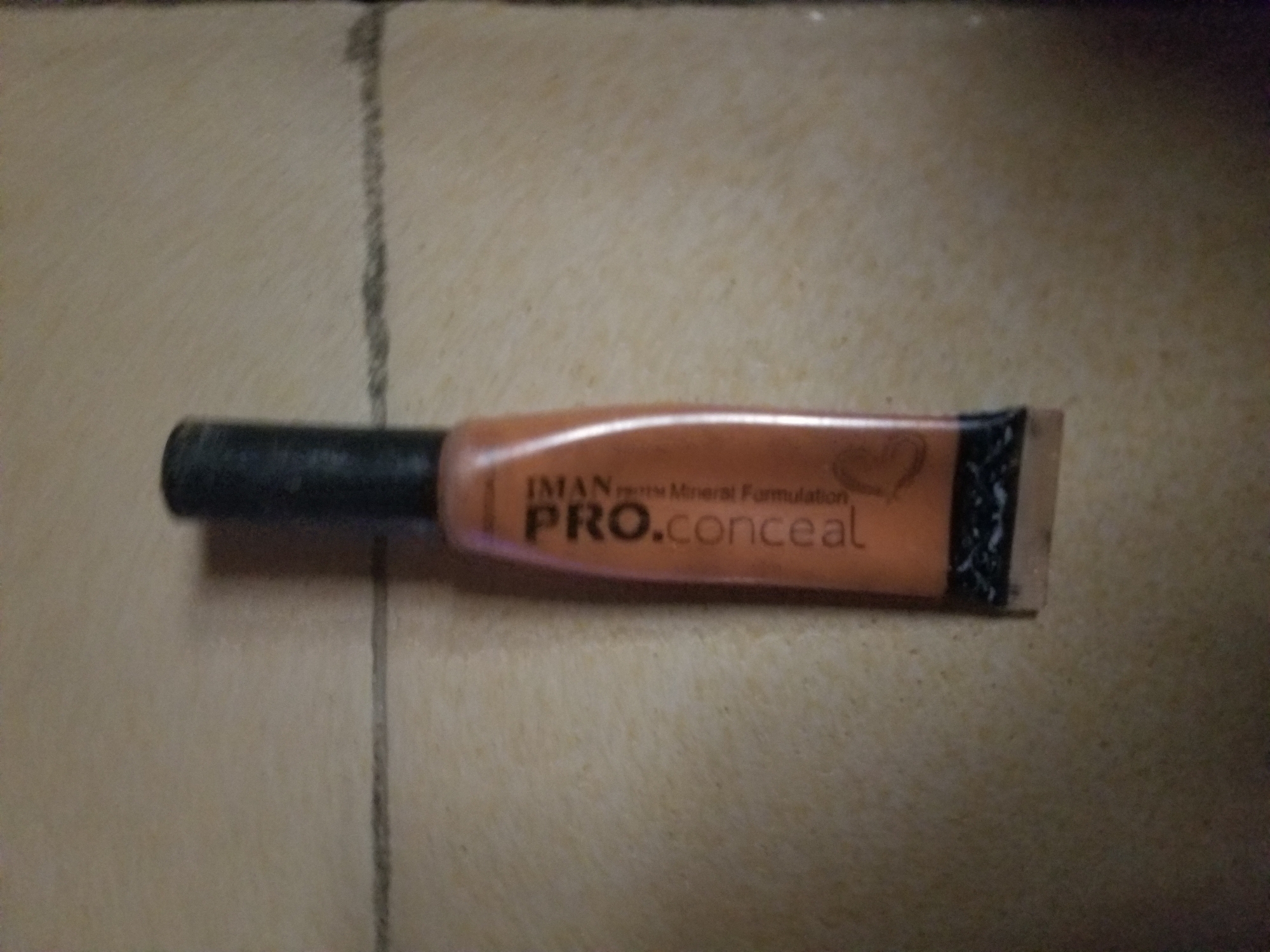 The image shows a tube of IMAN brand Pro.Conceal makeup. The tube is a light orange color and has black text on it. The text reads "IMAN pro Mineral Formulation PRO.conceal". There is also a small heart shape logo visible on the tube. The tube is lying horizontally on a textured surface that appears to be a floor or ground, with visible grout lines. The lighting suggests it might be indoors during the daytime. The product is an object and there is no activity or event taking place. The location context is Jauro Yinu, Nigeria.