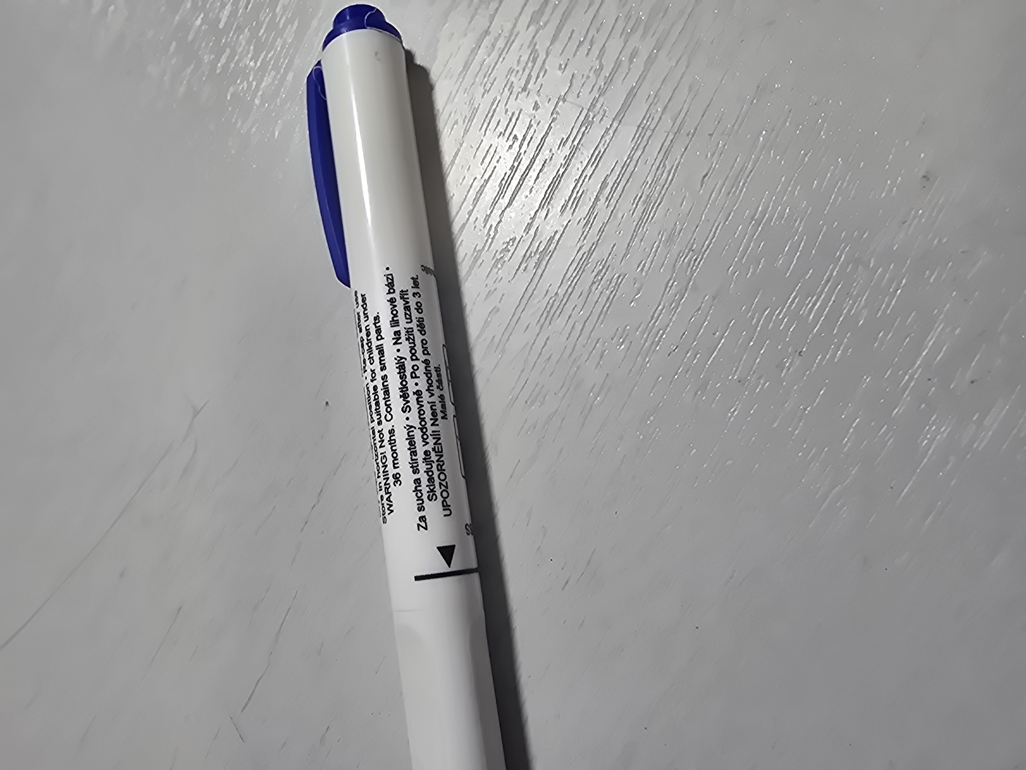 The image is a close-up, slightly angled, eye-level shot of a white dry-erase marker with a blue cap resting on a light-colored, textured surface. Text printed on the marker's barrel is partially visible, reading "Store in horizontal position • Re-cap after use WARNING! Not suitable for children under 36 months. Contains small parts." Below this, in what appears to be Czech, is "Za sucha stíratelný • Světlos... • Na lihové bázi • Skladujte vodorovně • Po použití uzavřít UPOZORNĚNÍ! Není vhodné pro děti do 3 let. Malé části." The surface appears to be wood with a visible grain pattern and some scuff marks. There are no people or animals present, and the background is out of focus, showing a general grayish-white tone. The scene is indoors and appears to be a tabletop. The location in Ulan-Ude, Russia cannot be determined from the provided image.
