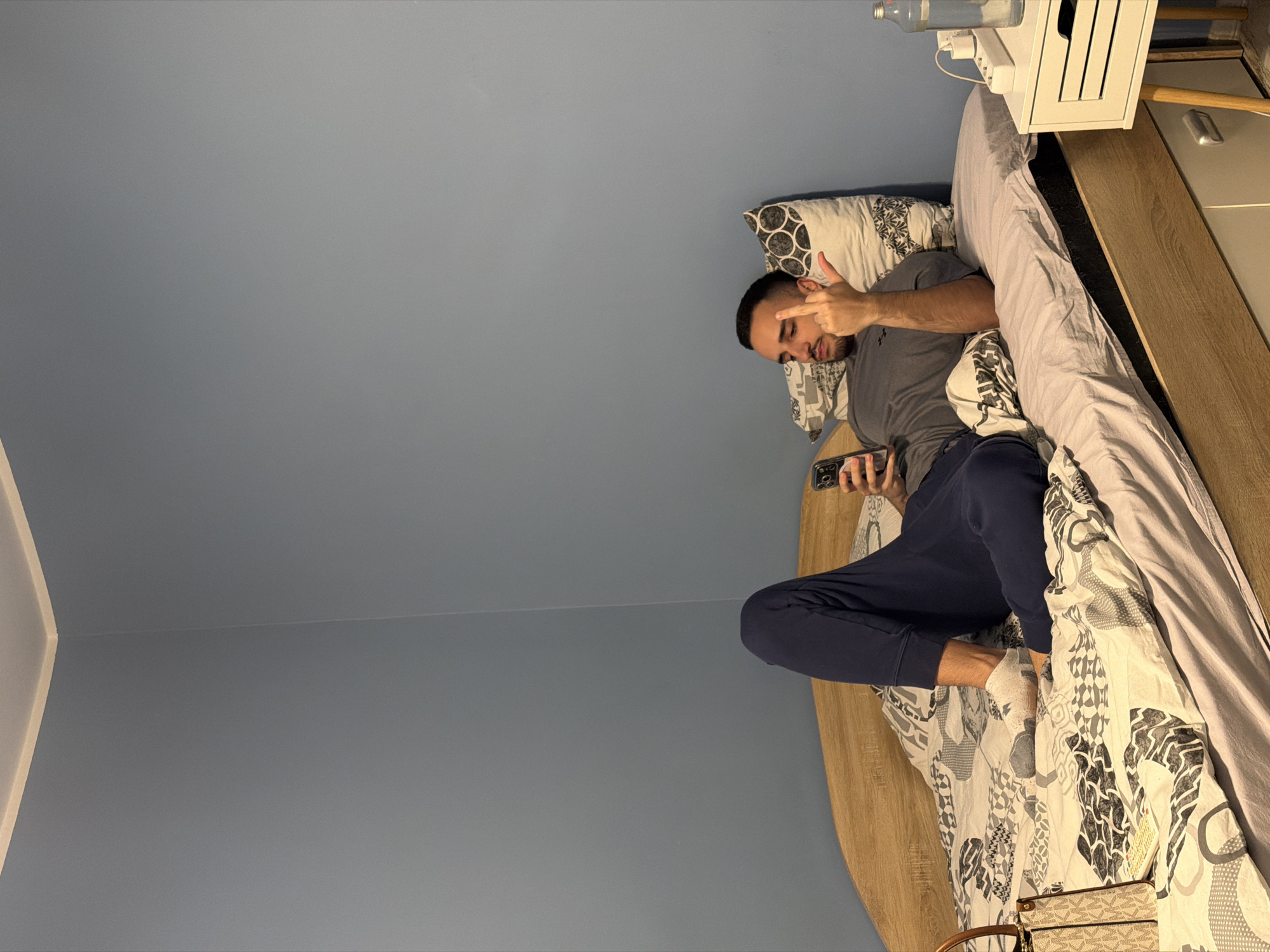An adult male is observed lying supine on a bed in a bedroom located in Plovdiv, Bulgaria. The room features medium blue painted walls, a light wood-toned bed frame, and a ceiling with a subtle light-colored trim. The bed is covered with a light grey fitted sheet and a duvet displaying a pattern of black, white, and grey abstract shapes. Two pillows support the individual's head, one matching the duvet pattern and another with a black and white circular design.
The male subject wears a grey t-shirt, dark blue sweatpants, and light grey socks. He holds a dark-colored smartphone in his left hand, visually engaged with the device. His right hand is raised, with the middle finger extended upward, forming a fist with the remaining fingers. To the left of the subject's lower body, a light brown Michael Kors monogrammed handbag rests on the duvet. A white two-drawer nightstand with wooden legs is positioned to the right of the bed. On the nightstand, a white power strip and a clear glass water bottle are visible.
