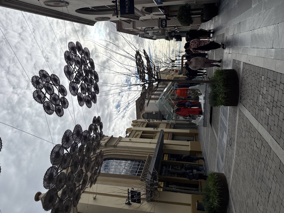 The image depicts a pedestrian shopping street within Las Rozas Village, located in Las Matas-pinar-monte Rozas, Spain, under an overcast sky. The street is lined with multi-story buildings featuring light-colored facades, arched windows, and traditional architectural details. Overhead, dark, circular, woven decorative elements are suspended by wires across the thoroughfare in multiple clusters.
The pavement consists of a pattern of light and dark grey rectangular and square pavers. Bare deciduous trees are planted in circular, dark-colored planters filled with green ground cover, positioned along the street.
On the left, the storefront for "COACH" is visible, with several individuals partially obscured near its entrance. In the mid-ground, a clear glass-enclosed display case occupies the center of the walkway, showcasing multiple mannequins dressed in winter apparel, including red jackets and patterned trousers, set on a base resembling snow.
On the right side of the street, storefronts for "HACKETT" and "UGG" are identifiable. Several people are present, walking along the paved street. In the foreground-midground, a woman in a light-colored top and dark pants walks from left to right, followed by a woman in a patterned top pushing a dark-colored stroller or cart. Additional pedestrians are visible further down the street, primarily dressed in casual winter clothing. No distinct interactions are observable beyond general movement.