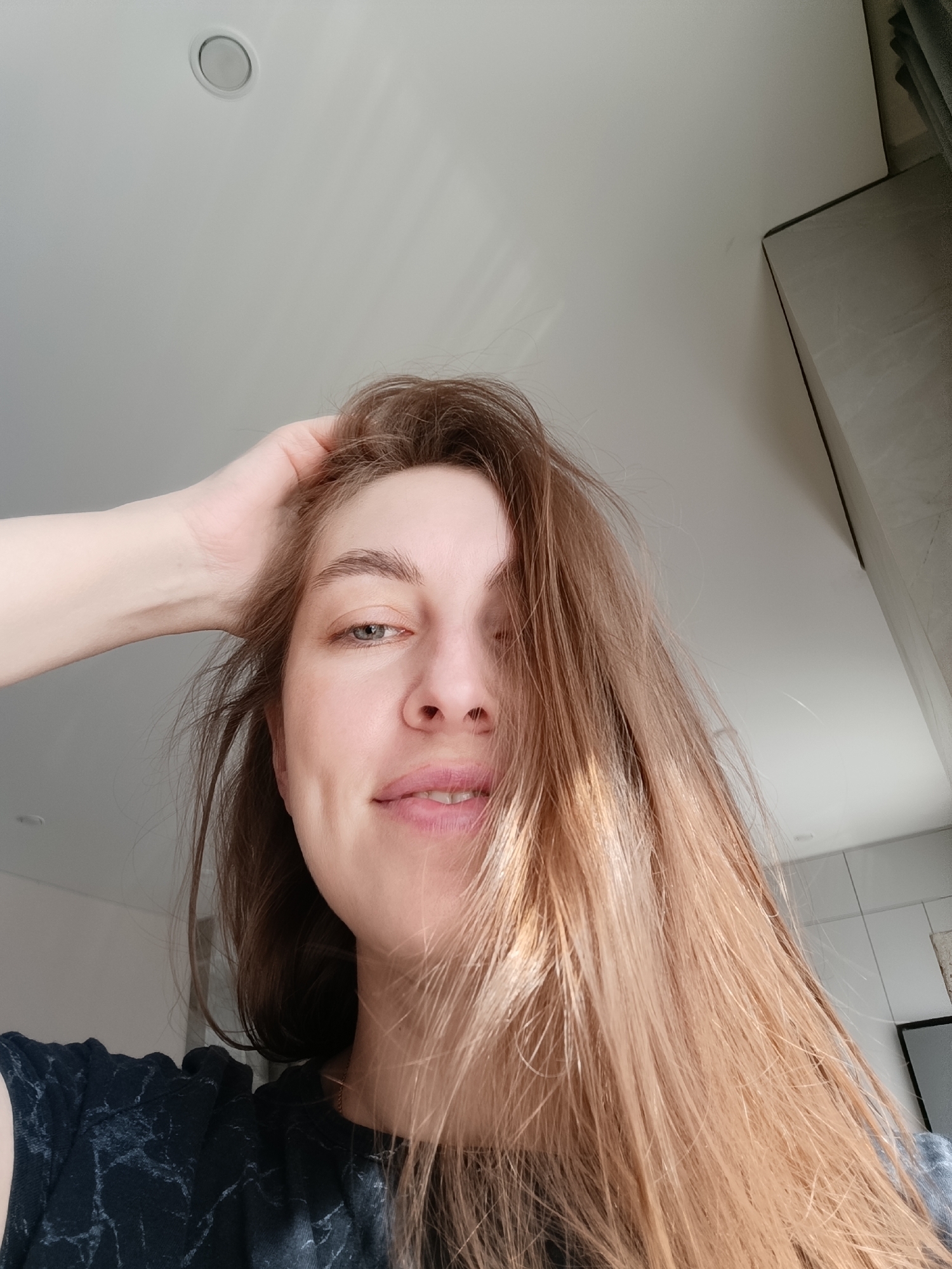 A fair-skinned woman with brown hair is facing the camera from a low angle. She has one hand in her hair. She is wearing a dark shirt with a marble-like pattern. The background is a white ceiling with a recessed light fixture and a column covered in a light grey material. The image appears to be taken indoors. There is no information in the image to determine that it was taken in the city of Chaikovsky, Russia.