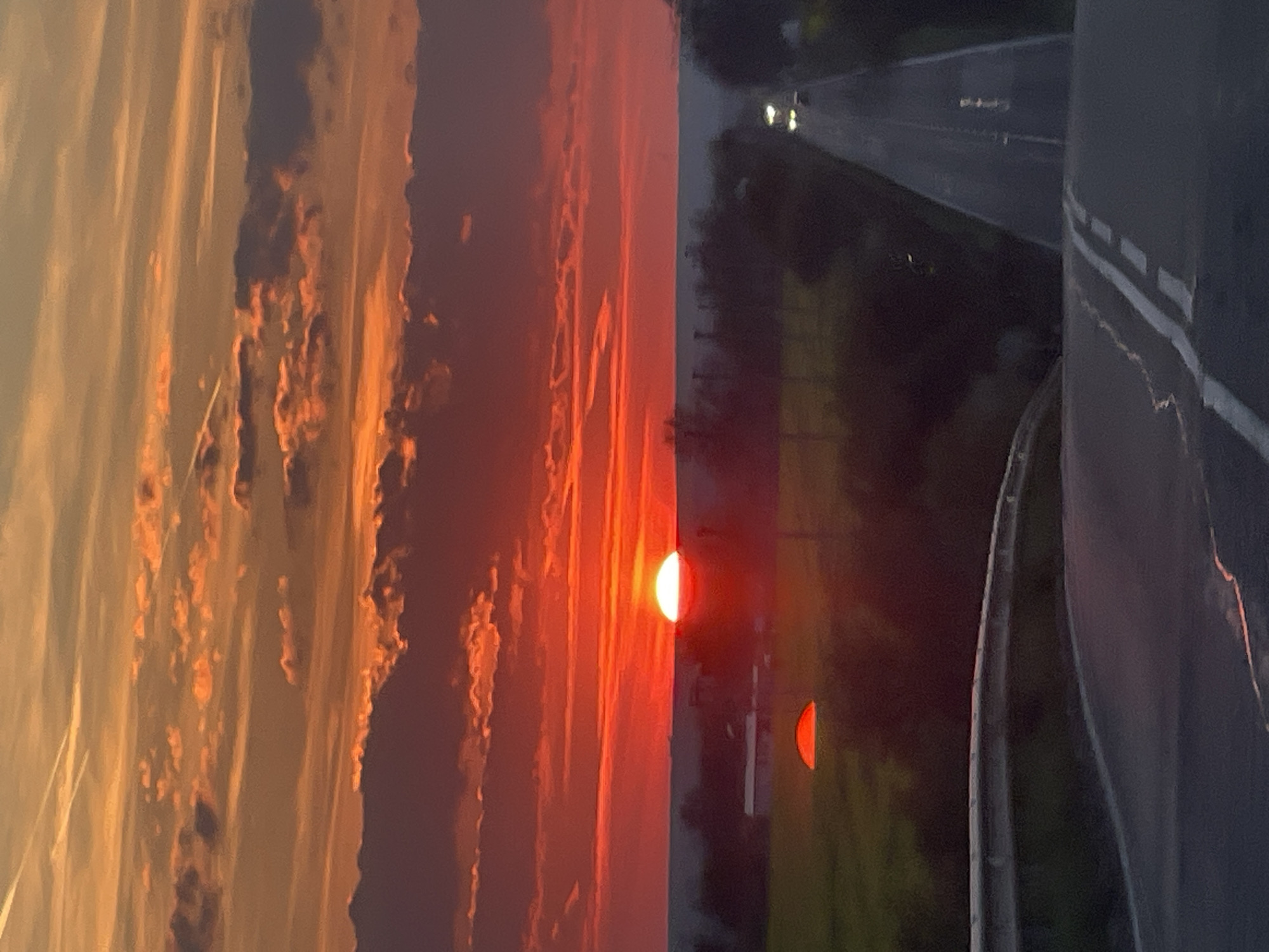 The image shows a sunset view adjacent to a road. The left side of the image is dominated by a vivid sunset featuring orange and red clouds. The sun is partially visible near the center, appearing as a red semi-circle. To the right of the sunset is a road, showing traffic with visible headlights. A guardrail is present along the road's edge. The image's location and the presence of the road and guardrail suggest the sunset is being viewed from an elevated position. The image's location, Bratovo, Bulgaria, is not immediately determinable from the content.
