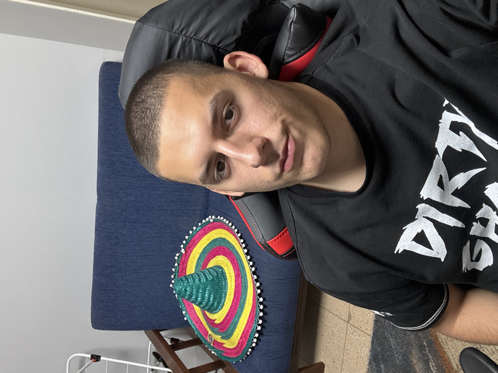 An upright-oriented indoor selfie features a young man with short, buzzed dark hair, seated in a black and red gaming chair. He looks directly at the camera with a calm, slight smile. He is wearing a black t-shirt with white text; the word "DIRTY" is clearly visible on his chest.
To his left, resting on a blue upholstered piece of furniture, is a vibrant sombrero. The sombrero has concentric rings of green, yellow, and pink, and its brim is decorated with small white pom-poms.
The background includes a plain light-colored wall and a floor with light tiles and a patterned rug. A white metal structure, possibly a drying rack, is partially visible in the lower left. The lighting is bright and even, indicating a daytime setting in a casual room, likely in Plovdiv, Bulgaria.