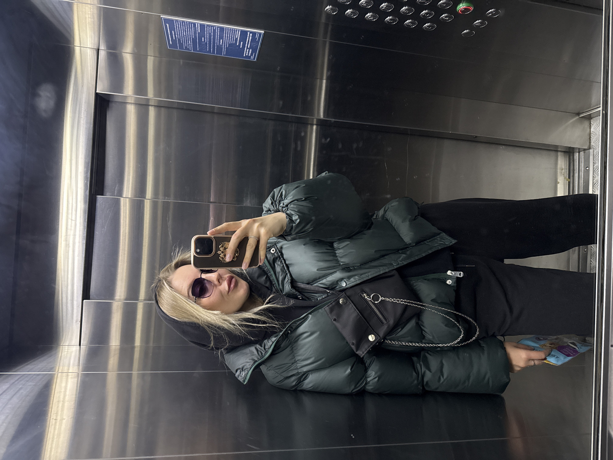 A woman is taking a selfie in an elevator. The elevator's interior is stainless steel with a panel of buttons at the top. The woman is wearing sunglasses, a hooded sweatshirt, a dark green puffer jacket, and dark pants. She holds a smartphone in her right hand and a blue package in her left. A small black purse hangs from her jacket. A blue information plaque is attached to the upper wall of the elevator. The scene likely takes place in Krasnoyarsk, Russia.