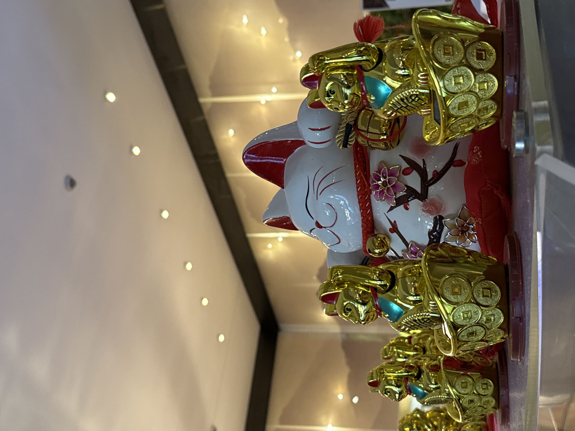 The image depicts a collection of golden maneki-neko figurines arranged on a shelf, with a larger white and red maneki-neko positioned centrally behind them. The figurines are in various sizes and appear to be made of a reflective gold-colored material. The smaller figurines are facing left and are sitting in golden bowls adorned with coin motifs. The larger central figurine is predominantly white with red accents on its ears, collar, and facial markings, and it also holds a golden object. The background features a warm, diffused light with several circular light fixtures visible on the ceiling. The overall scene suggests an indoor display, possibly in a shop or home. The provided metadata indicates the location as Nordhausen, Germany, but this cannot be visually confirmed from the image itself.