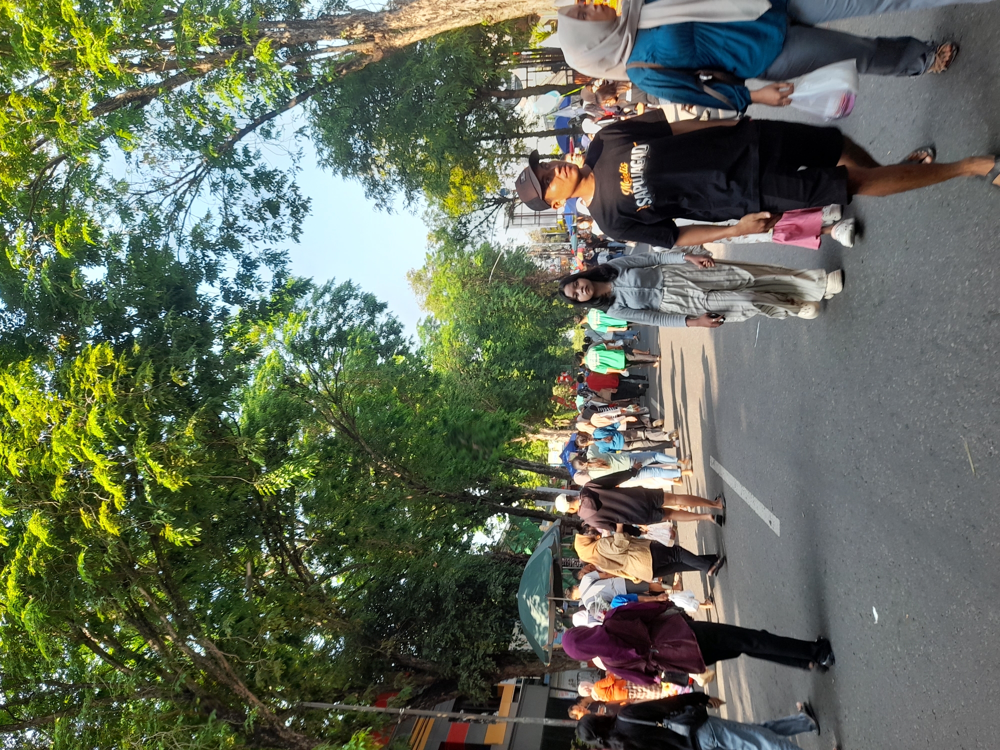 In Purwodadi, Indonesia, a street is lined with mature trees casting dappled shade on a moderately busy scene. The paved road, marked with a white dashed line, is occupied by a diverse crowd of people walking in various directions.
In the foreground, a young man wearing a black t-shirt with "Majelis SAPUJAGAD" printed on it, black shorts, a black baseball cap, and sandals walks towards the viewer. To his left, a young woman in a light gray long-sleeved top and striped pants also walks towards the viewer. To the right of the foreground male, a woman with a light gray hijab and a teal top is visible.
Further back, a woman in a maroon hijab and black pants is walking away from the viewer. Other individuals are seen in the middle ground, some carrying shopping bags. There are several market stalls or vendor tents visible along the sides of the road, with green canopies. The buildings on the left side of the street appear to be commercial establishments. The sky is clear and bright blue, suggesting a sunny day. The overall atmosphere appears to be that of a lively street market or public gathering.