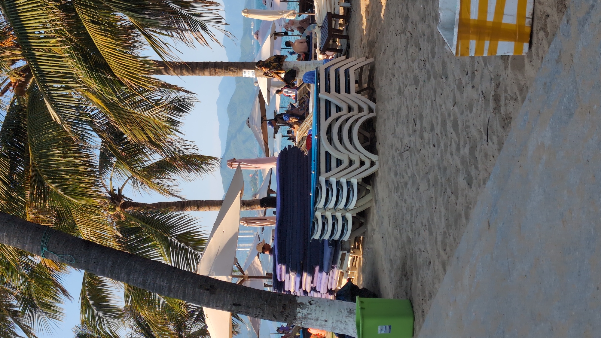 This image depicts a beach scene in Nha Trang Ward, Vietnam. In the foreground, there is a pile of stacked blue lounge chair cushions on top of stacked empty lounge chairs. To the left, a green trash bin is visible. Palm trees with green fronds arch over the scene, with their trunks in the foreground and midground. In the background, several beach umbrellas and lounge chairs are set up on the sand. People are visible sitting on lounge chairs and walking near the water. The sea stretches out to the horizon, with hills or mountains in the distance. The sky is clear and blue. The overall impression is that of a relaxed beach atmosphere.