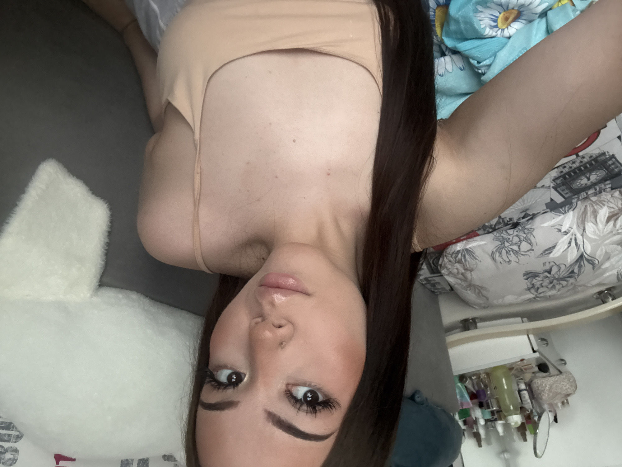 A young woman lies on a bed in an indoor setting, with a white pillow to her left and patterned bedding on her right. She is wearing a beige top, and her dark hair is neatly styled. She appears to be looking directly at the camera. In the background to her right, there is a shelf with various bottles and cosmetic items. This image was taken in Naberezhnye Chelny, Russia.