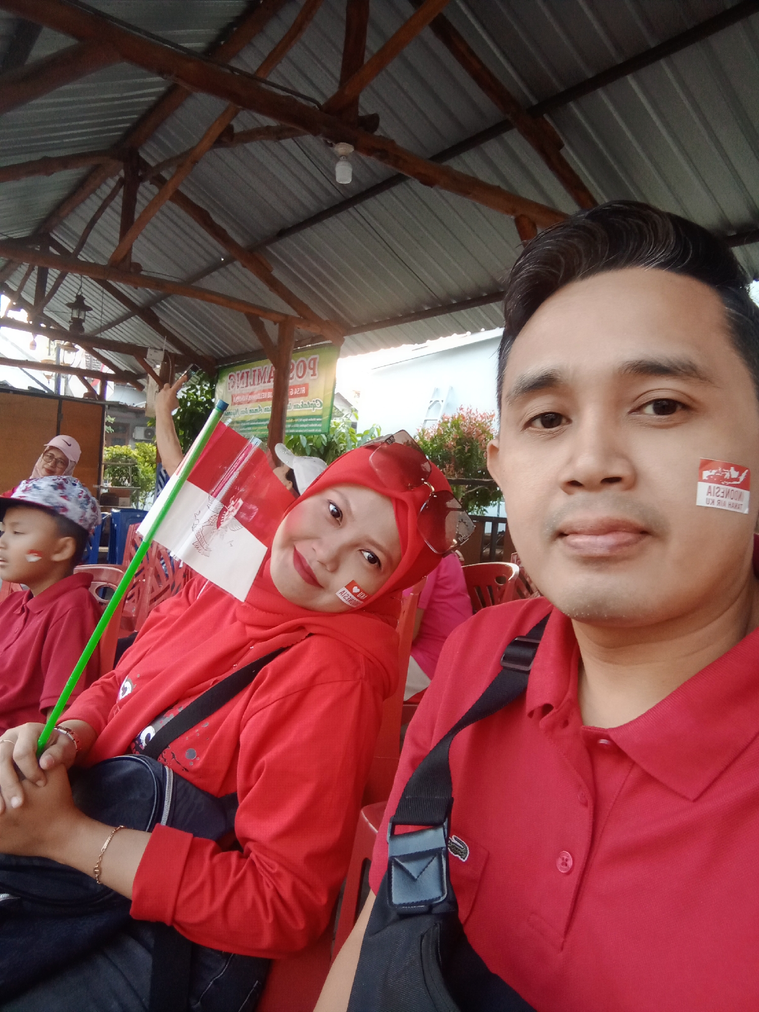 A man and a woman, both wearing red shirts and sporting Indonesian flags on their cheeks, pose for a selfie under a wooden and corrugated metal roof. The woman, in the foreground, is holding a small Indonesian flag. In the background, a young boy in a red shirt and a patterned cap is visible, as well as another woman wearing glasses. The scene appears to be an outdoor gathering or event. The man in the foreground has a black strap from a bag across his chest. A white and green sign with text in Indonesian is visible in the background. The overall impression is one of patriotic celebration.