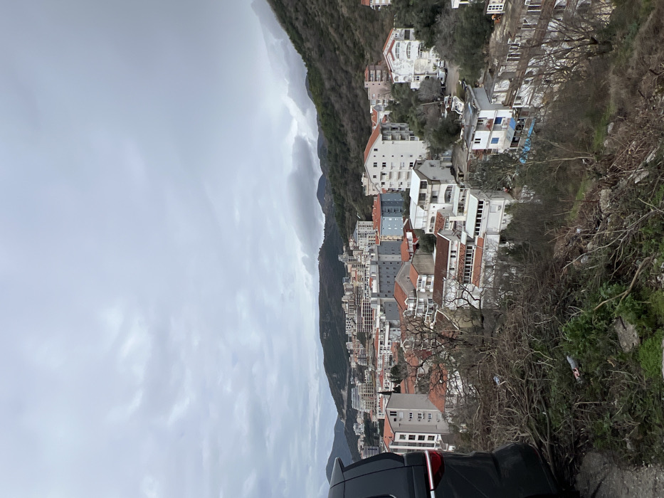 This image presents a panoramic view of the city of Rafailovići, Montenegro, situated on a hillside under a heavily overcast sky. In the immediate foreground, to the left, the rear quarter panel and partial roof of a dark-colored vehicle, possibly an SUV or minivan, are visible. The vehicle displays a vertical red taillight element. The foreground terrain consists of a steep, unkempt slope covered with dry brush, scattered cut branches, and sparse green vegetation.
The midground features a dense concentration of multi-story buildings, characteristic of a coastal town. These structures exhibit varied architectural styles, including buildings with prominent red-tiled roofs and others with flat or light-colored roofs. Building facades range from white and light grey to blue and exposed concrete, suggesting a mix of residential and commercial properties. A winding road or open area with several parked white vehicles (likely vans or cars) is visible in the lower-right midground, amidst the buildings. Green trees and other vegetation are interspersed throughout the urban landscape.
In the background, a series of rolling, forested mountains frame the city, extending towards a hazy horizon under the heavy grey cloud cover. No individuals are discernible in the scene.