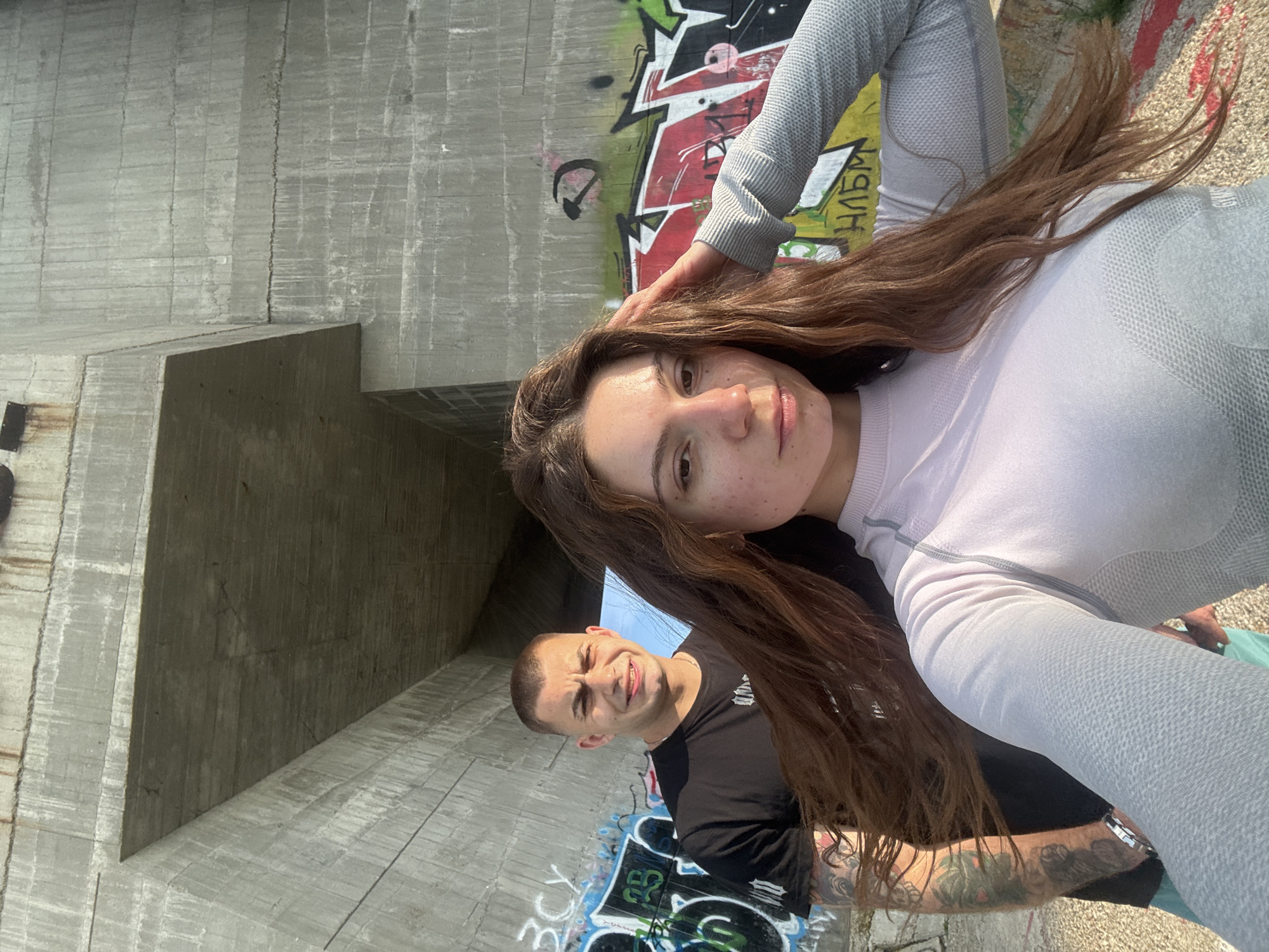Two individuals are in a selfie-style photo taken outdoors. A woman with long brown hair is in the foreground, wearing a long-sleeved, light-colored top. Her expression is neutral. A man with short, dark hair, wearing a black shirt, is slightly behind her and to the left. He appears to be smiling.
The background consists of a large concrete structure, possibly a bridge support or wall, with graffiti visible on the surface. The concrete structure appears to be aged with weathering. The location could be in the city of Varna, Bulgaria. The angle of the photo suggests they are either standing or crouching, holding the camera at an upward angle.