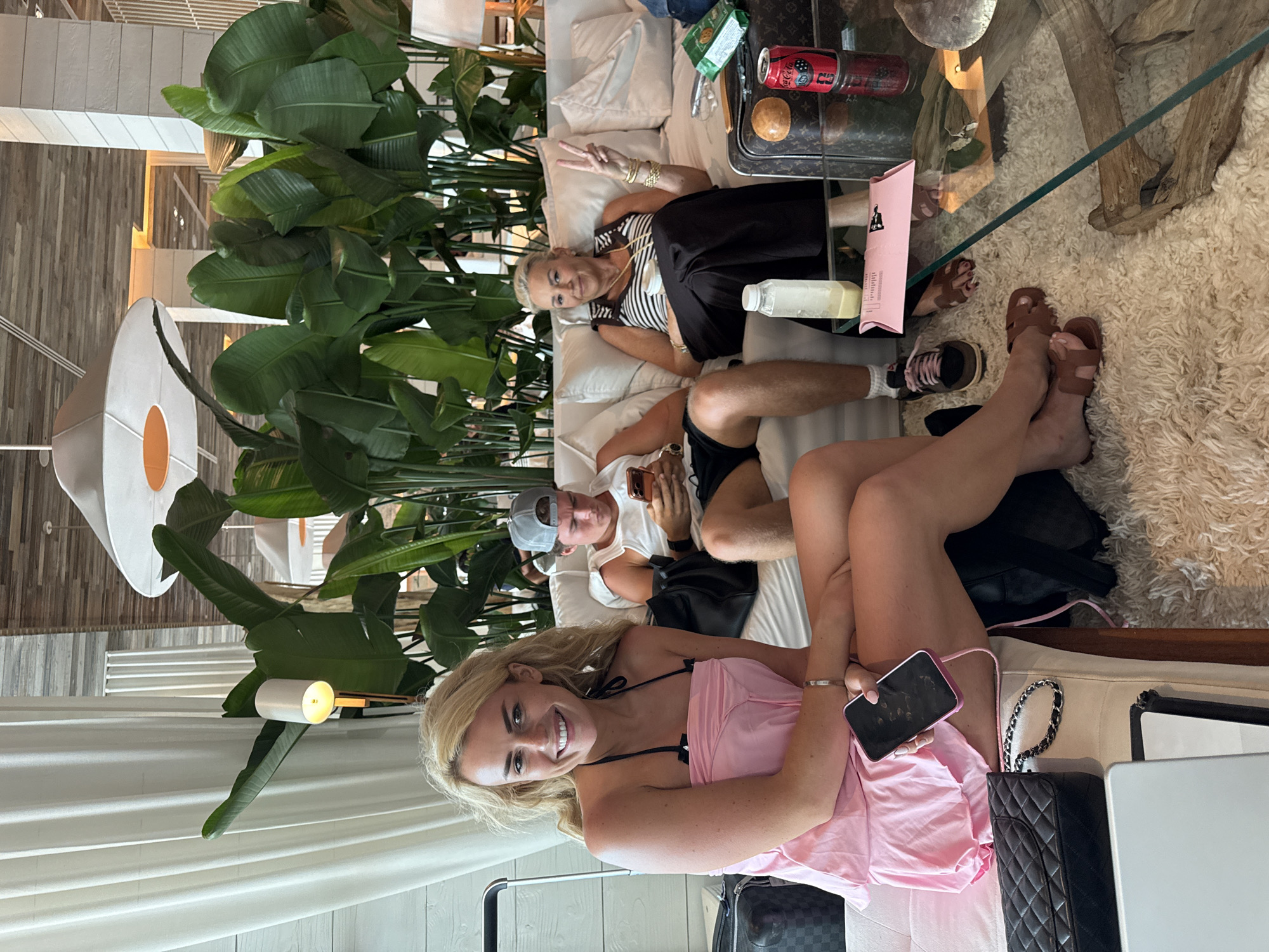 This media file depicts three individuals relaxing in a modern, plant-filled indoor lounge, likely in Miami Beach, United States of America. The overall atmosphere suggests a casual, leisure activity.
In the foreground, a young woman with blonde hair, wearing a pink camisole and shorts, smiles directly at the camera while holding a black smartphone. Her bare legs are visible, and a quilted black handbag rests beside her. Behind her, a man wearing a white t-shirt and a backward baseball cap is reclined on a light-colored sofa, looking at his own smartphone. Further back on the same sofa, an older woman with blonde hair, dressed in a striped top and black skirt, smiles and makes a peace sign with her hand.
The setting is an elegantly furnished lounge area. Large, vibrant tropical plants (possibly Bird of Paradise) form a lush backdrop behind the sofa. A clear glass coffee table stands on a fluffy, cream-colored shag rug. On the table, a red Coca-Cola can, a white bottle, and a small pink paper bag with some visible text are present, along with what appears to be a Louis Vuitton pattern on an item. The floor in the background features wooden slat panels. A modern white light fixture with an orange circular accent is visible on the left. Other details include several cell phones, brown sandals, and a visible suitcase handle.
The scene conveys a relaxed and pleasant mood, with two of the subjects smiling. The lighting suggests it is daytime. The modern decor, tropical plants, and casual attire are consistent with a resort or hotel lounge in a vacation destination like Miami Beach.