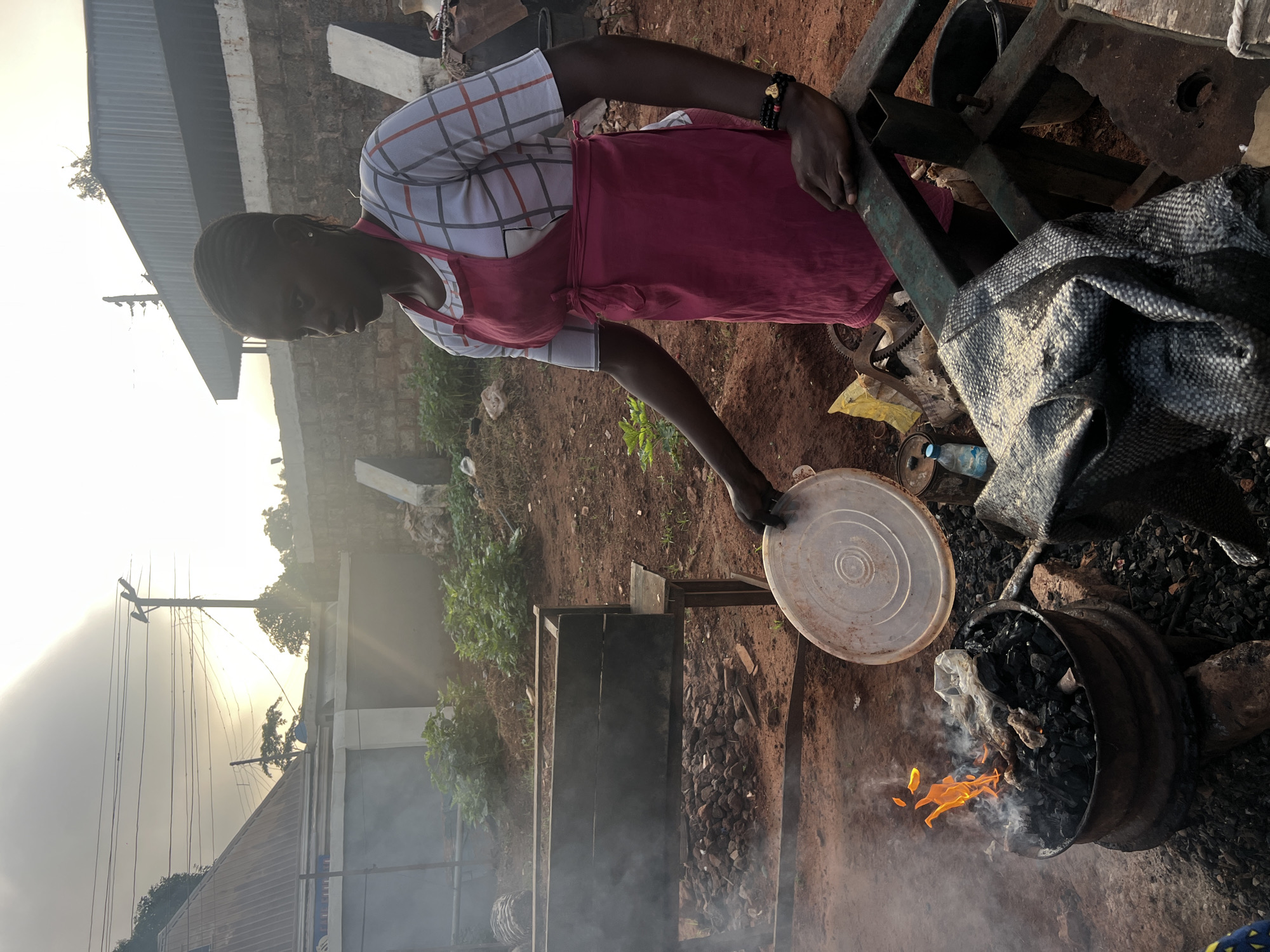 In Nkpor, Nigeria, a young woman wearing a pink apron over a checkered shirt is tending to a fire. The fire, fueled by charcoal, burns in a metal basin, producing smoke that drifts into the hazy outdoor environment. She holds a plastic lid in her right hand, and her left arm rests on a metal structure. The ground is dirt, littered with rocks and debris. In the background, a long wooden structure sits on legs, possibly used for cooking or display. Beyond that, there are buildings with corrugated metal roofs and a brick wall. The sky is bright with sunlight, suggesting it is either early morning or late afternoon. The overall scene depicts a street vendor or cook engaged in outdoor food preparation.