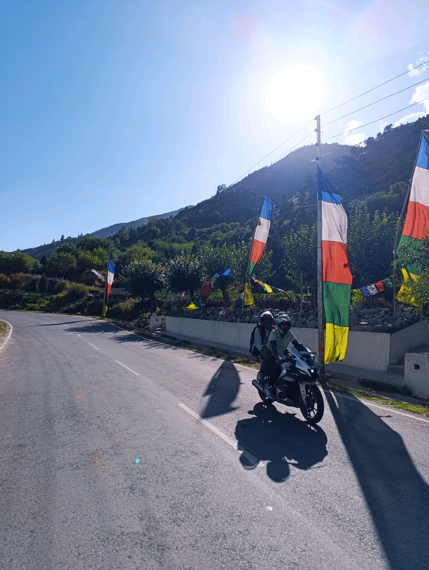 In Gemur, India, a motorcycle carries two individuals along a paved road, casting long shadows in the bright sunlight. The motorcycle is silver and white, and both riders wear helmets. Prayer flags in red, white, blue, green, and yellow are strung on poles, fluttering in the breeze, adjacent to a low stone wall. Behind the wall, a hillside covered in trees and rocks rises towards a clear blue sky, with the sun positioned high and to the left, creating a lens flare. The road curves to the left, disappearing into the distance.
