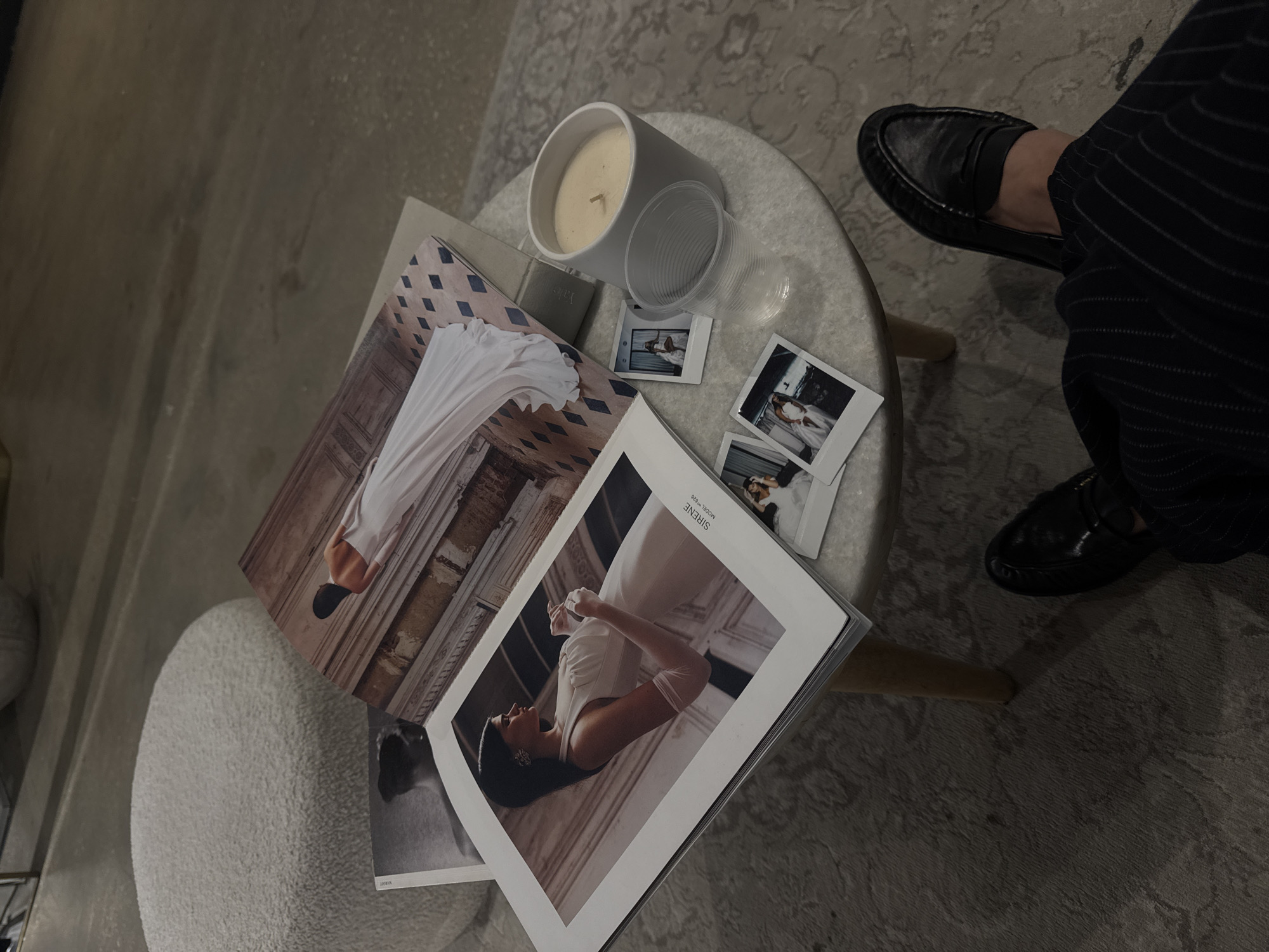 The image captures a vignette of a stylish, personal space. A round, light-colored table hosts a curated collection of items: a lit candle in a minimalist grey holder, a clear plastic cup, a small grey box, and a stack of printed photographs and open magazines or books. The magazines appear to feature fashion photography, specifically brides and wedding dresses, with images of models in elegant gowns. The photographs seem to be snapshots of the same subject matter.
The edge of a white, textured stool is visible on the left side of the frame. To the right, the lower portion of a person is seen wearing striped pants and black leather loafers.
The location is unlikely to be identifiable as Tel-Aviv, Israel, from the visual information provided.