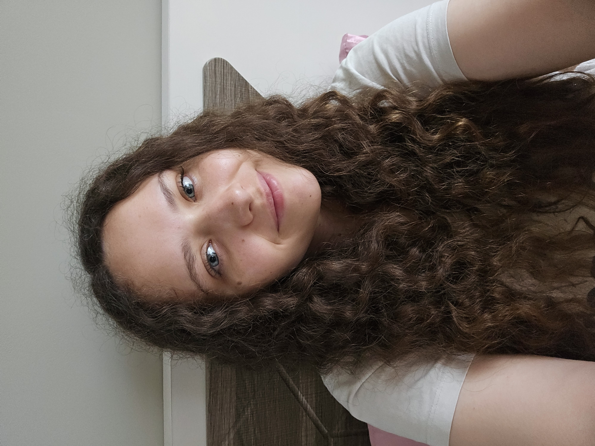 This is a close-up, selfie-style image of a young person with long, curly, dark brown hair. They are lying down and looking up towards the camera, with their head tilted. The image is oriented vertically, so the person appears sideways.
The subject has striking light blue eyes with flecks of gray. Their skin is fair with a few freckles visible on their cheeks and nose. They have a gentle, slightly closed-mouth smile, suggesting a relaxed or content emotion. The lighting appears to be indoor, with a soft, diffused light source, potentially from a window or artificial light, creating subtle shadows on their face.
They are wearing a plain white t-shirt. Behind them, a light gray wall is visible, along with a portion of a wooden surface that appears to be part of a piece of furniture, possibly a headboard or shelf, with a textured, grainy pattern. The overall setting seems to be a bedroom or a similar indoor private space.
There is no visible text in the image. The time of day and weather are not discernible from the visual information. The image conveys a sense of intimacy and casualness, typical of a personal selfie.
