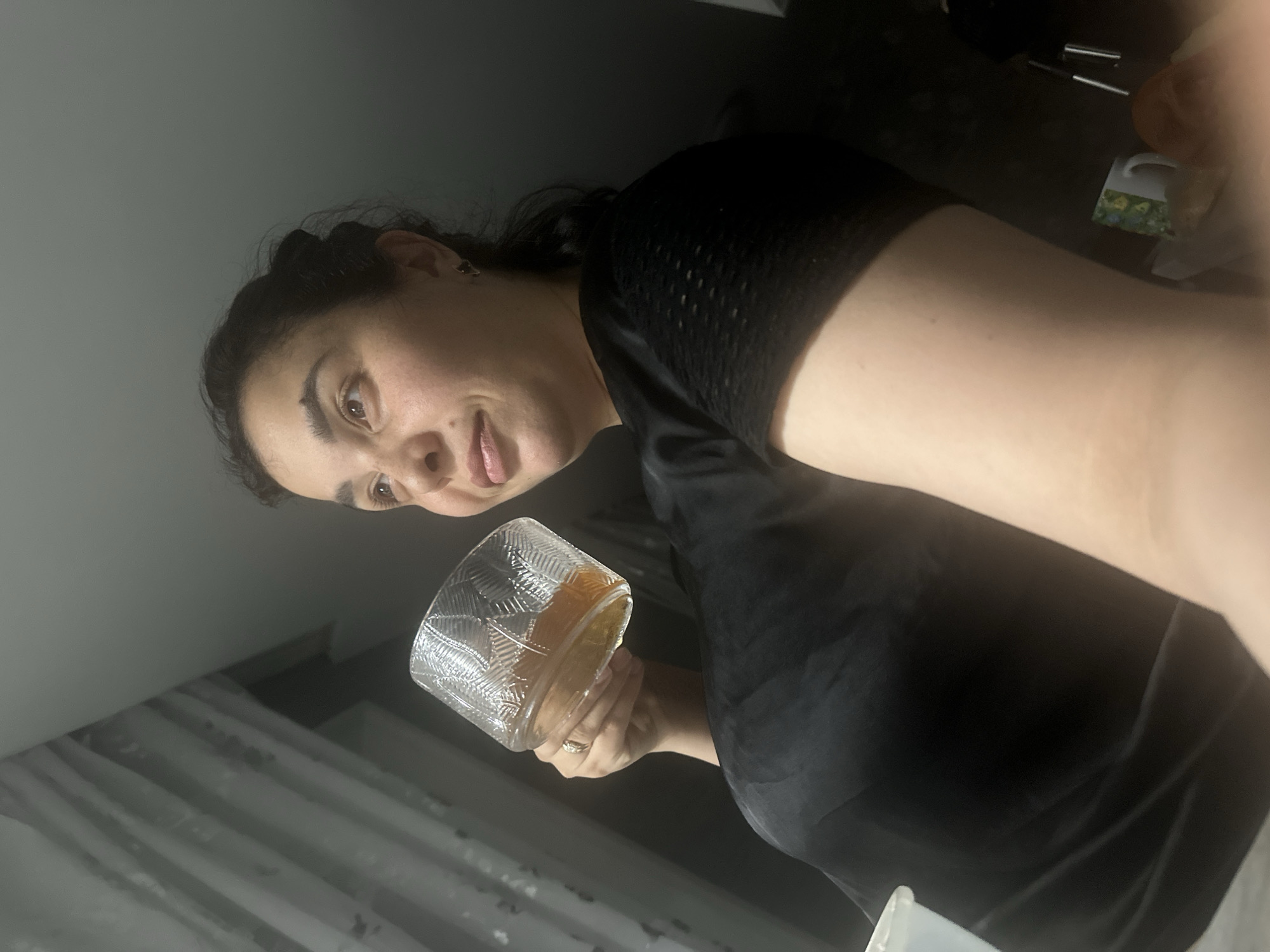 A woman is holding a patterned glass containing a yellow liquid. She is wearing a black shirt. The background is dimly lit with a grey wall and what appears to be a sheer curtain. The location is Unknown, Unknown.