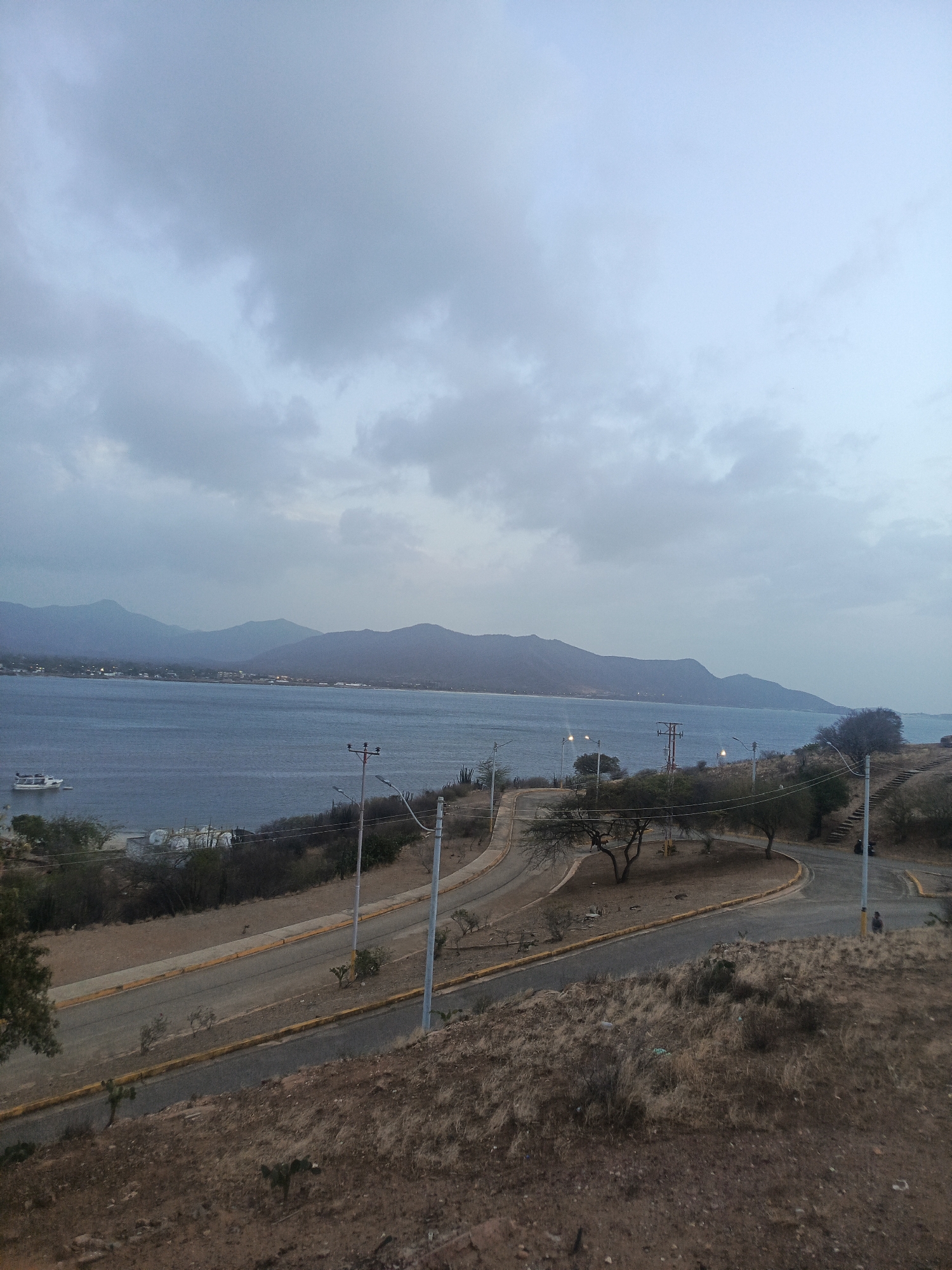 This wide-angle photograph presents an overcast coastal landscape near Juan Griego, Venezuela, under a sky dominated by soft grey clouds. A tranquil, blue-grey body of water occupies the middle ground, framed by a series of distant, undulating mountains that stretch along the horizon. A white boat or yacht is visibly anchored or docked in the water on the left side of the frame.
In the foreground, a dry, arid hillside covered in brownish grasses, scrub brush, and scattered small trees slopes down towards the coast. Paved roads with distinct yellow curbs wind through this terrain, suggesting a developed area. Several utility poles with streetlights are positioned along these roads, with some power lines also visible. On the right, steps are carved into the hillside. Two small, indistinct human figures are discernible on the roads; one near the upper right and another further down the road below the steps.
The diffused, muted light indicates either an early morning, a late afternoon, or simply a very cloudy day, contributing to a calm and somewhat subdued atmosphere. The general appearance of the vegetation, coastal mountains, and arid landscape aligns with the characteristics of Juan Griego on Margarita Island. No visible text is present in the image.