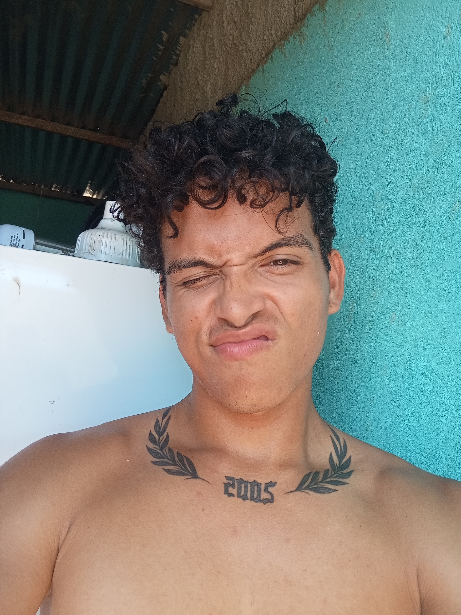 A young man with curly dark hair and tattoos on his chest stands in front of a teal-colored wall. He is making a humorous face, with his eyes squinted and mouth puckered. The background shows a corrugated metal roof and a white appliance. The scene appears to be outdoors, possibly in a residential area of Altagracia, Venezuela. The man has a tattoo of a laurel wreath on his chest, with the numbers "2005" beneath it. The overall impression is casual and playful.