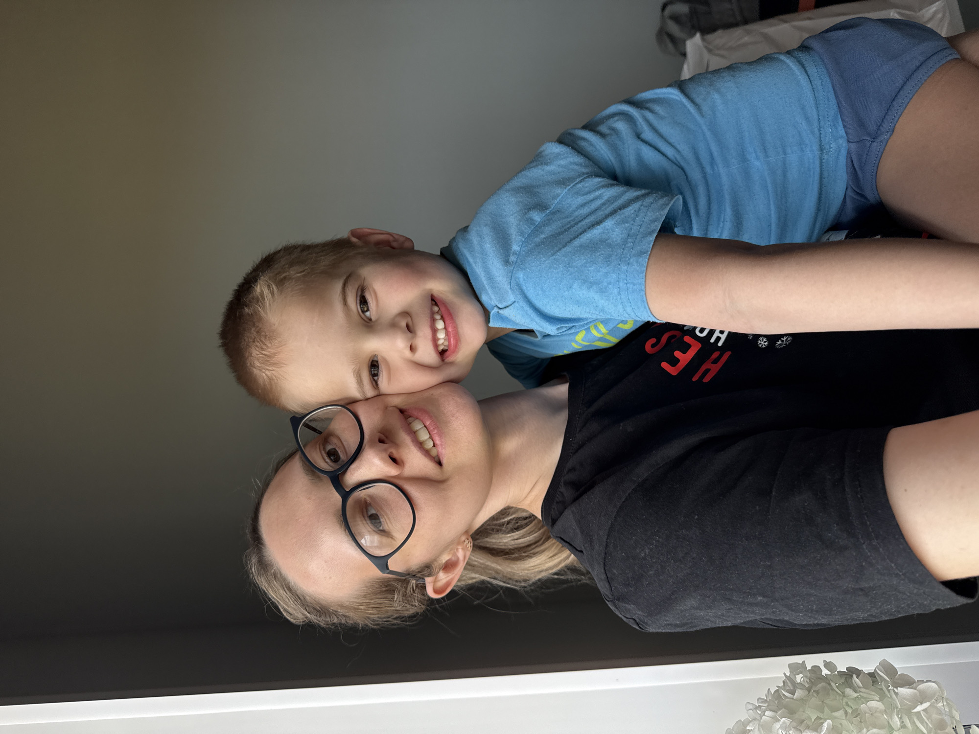 A woman and a young boy are posing for a selfie indoors. The woman is wearing glasses and a black shirt. The boy is on top of her, wearing a blue shirt and shorts. They are both smiling at the camera. The background is a plain wall and a window sill with a white object. The location is potentially Chaikovsky, Russia, but there is no visual confirmation of this.