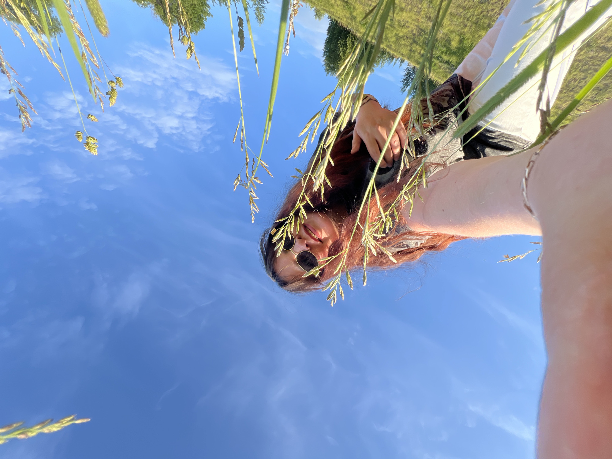 A low-angle selfie depicts a young female looking into the camera from a grassy field in the city of Noviy, Russia, under a clear blue sky with scattered white clouds. The camera is positioned low, capturing tall, green and dried grass stems with seed heads prominently in the foreground and midground. The female, with long reddish-brown hair, is wearing dark, round sunglasses with gold-toned frames and is smiling, revealing teeth. Her right arm is extended towards the camera, with a silver chain bracelet visible on her wrist, partially obscuring the bottom left of the frame. Her left hand is partially visible near her chest. She is wearing a dark top, possibly over a light-colored garment. In the background, distant trees and a grassy horizon are visible on the right, suggesting an open, sunlit landscape.