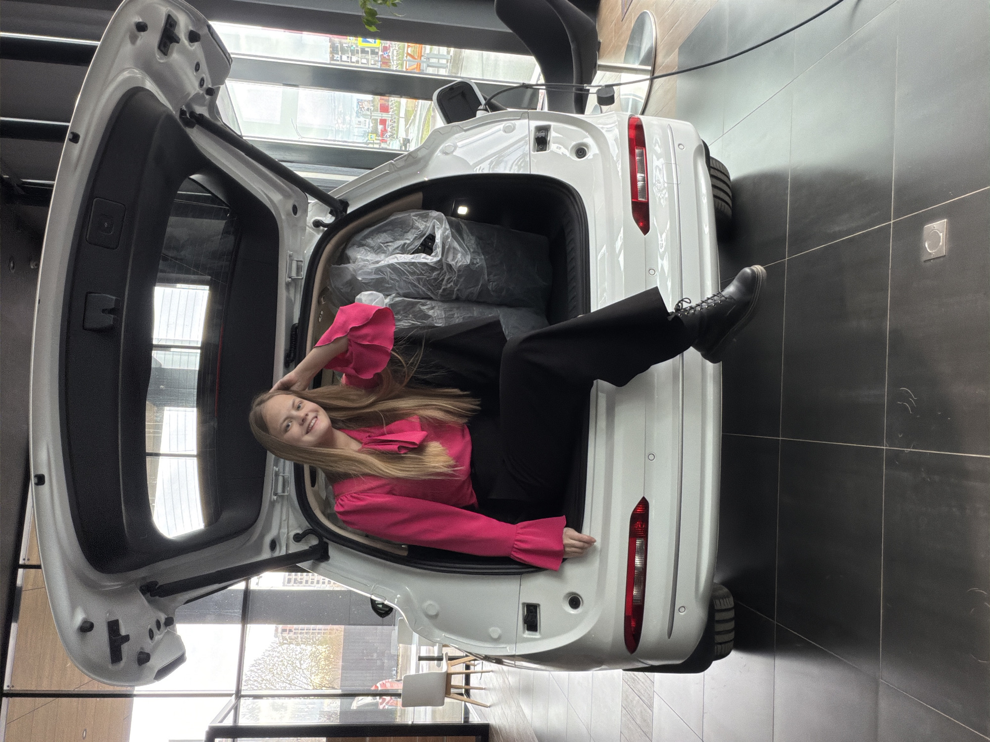 A young, fair-skinned woman with long blonde hair is lying in the open trunk of a white vehicle, presumably a car, in an interior space that looks like a showroom. She is wearing a pink long-sleeved blouse and black pants, and her left foot, clad in a black boot, is extending out of the car. The trunk is partially filled with objects covered in plastic wrap. The car's trunk lid is raised and open, revealing the interior of the car and also a partial view of an exterior cityscape through the windows in the background. In the background, one can discern details of a city with buildings and trees, possibly Izhevsk, Russia. The floor has dark tiles.