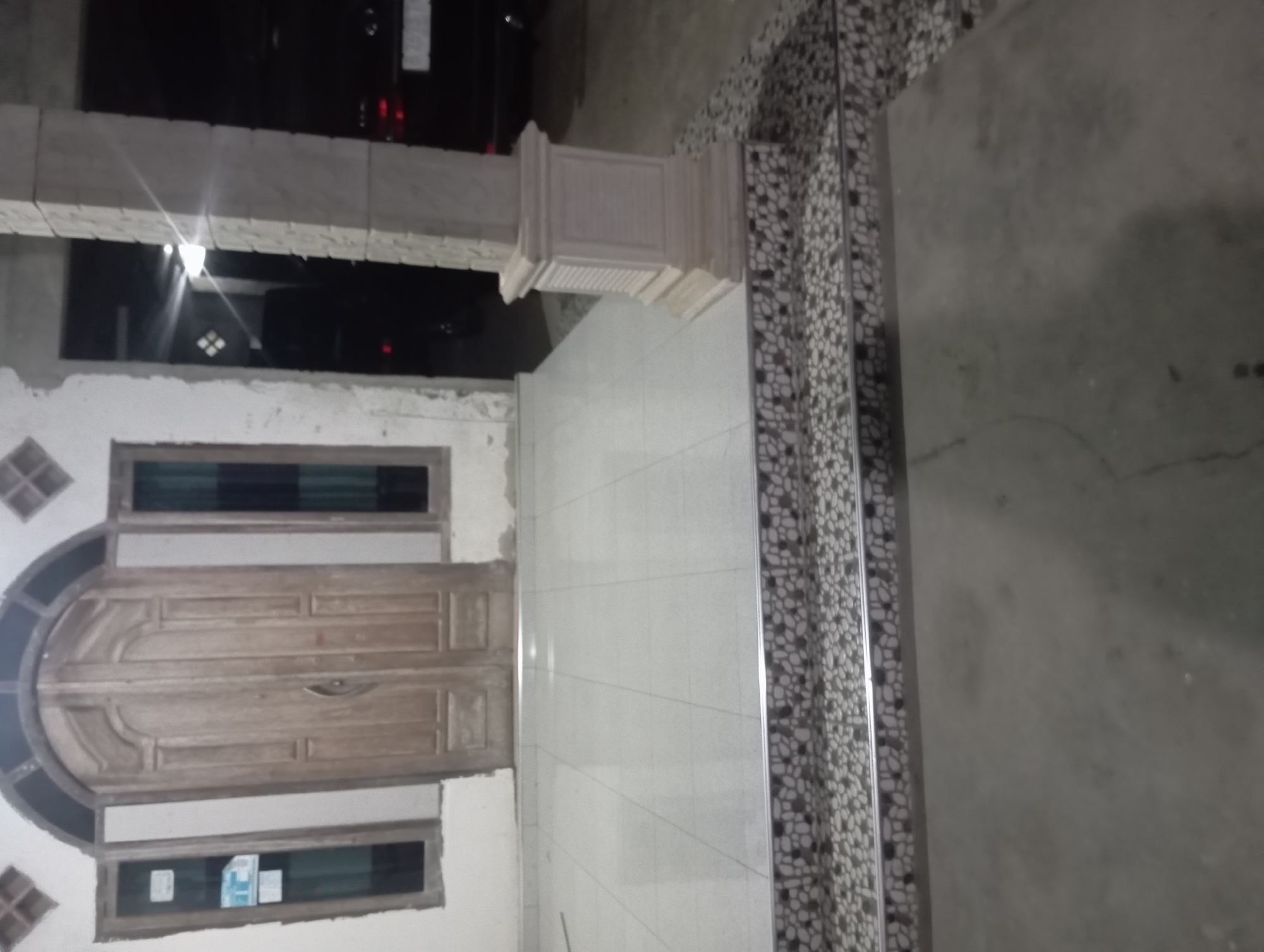 The image shows the exterior of a building in Rejosari, Indonesia, likely a residential home, at night. The foreground features a concrete ground leading up to a tiled walkway or porch. The walkway is bordered by a step with decorative stone-like tiles and a metal trim. To the left, a set of double wooden doors with arched detailing is visible, flanked by two tall, narrow windows with dark frames. Above the door and windows, the wall is a neutral color, with diamond-shaped decorative elements on the upper portion. To the right, a thick, decorative concrete pillar stands prominently, also featuring a layered, stepped design at its base. Beyond the pillar, a dark-colored vehicle, possibly an SUV or sedan, is partially visible, with its taillights faintly illuminated. The overall scene suggests a dimly lit, outdoor environment, with artificial light sources casting some illumination on the surfaces.