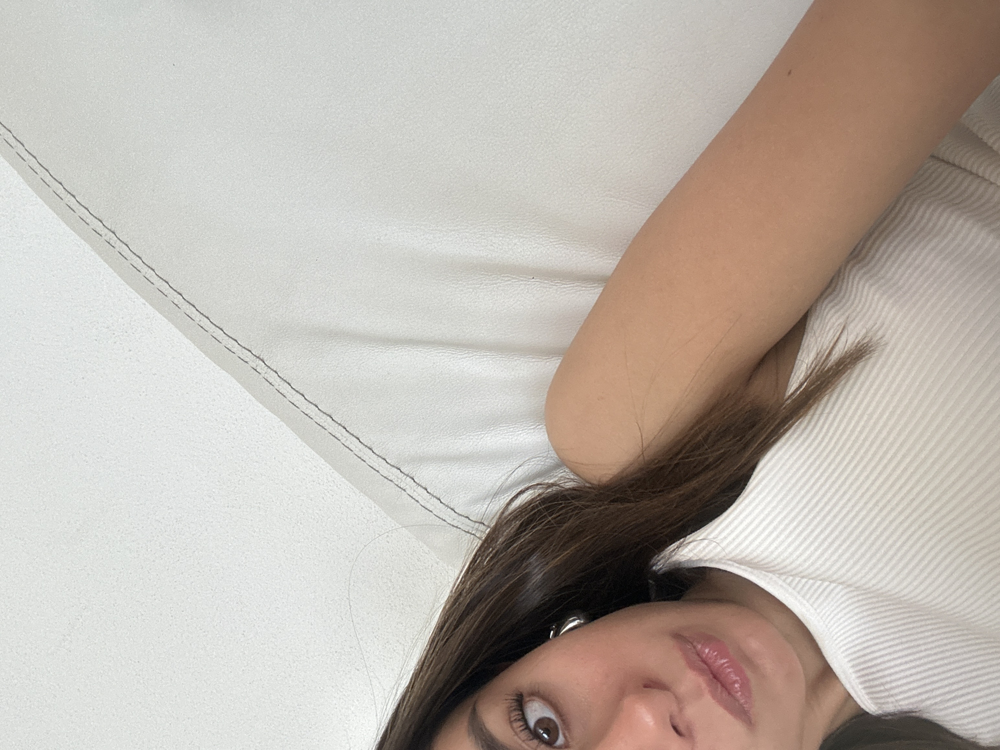 A close-up shot features a portion of a person's face, specifically the nose, lips, and left eye. The person has dark brown hair and is wearing a white ribbed top. The skin tone is fair. The background consists of a white, textured surface, likely a couch. The overall setting appears to be indoors. The city and specific location within the city are unknown.