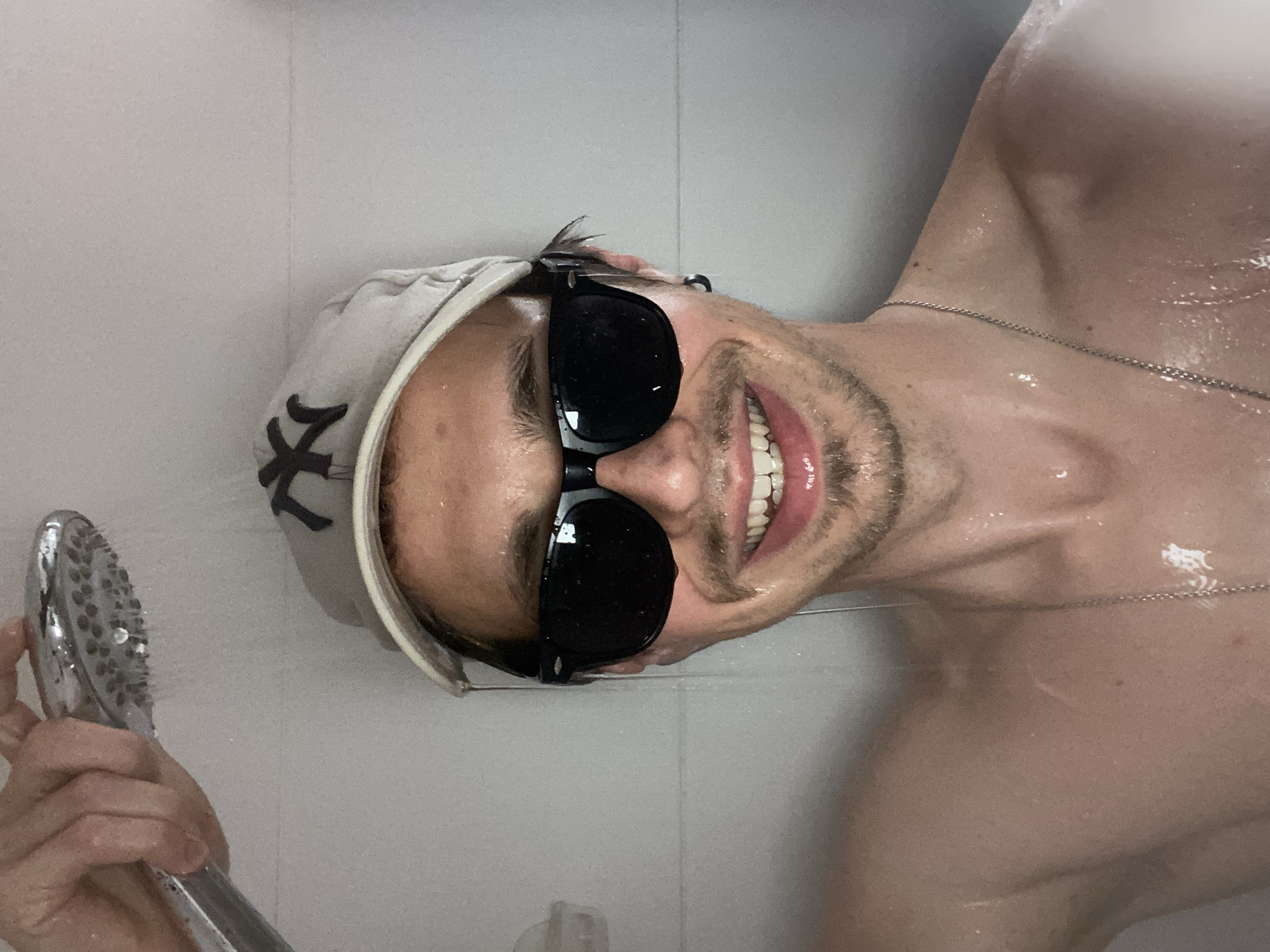 A light-skinned man with short brown hair, a trimmed beard, and wearing dark sunglasses and a light-colored baseball cap with a dark logo (possibly a stylized "NY") is in a shower. He is holding the showerhead in his hand, with water flowing from it. He is wearing a thin silver chain around his neck. The shower has a gray tile wall. There is no information present in the image to determine the location as Šušanj, Montenegro.