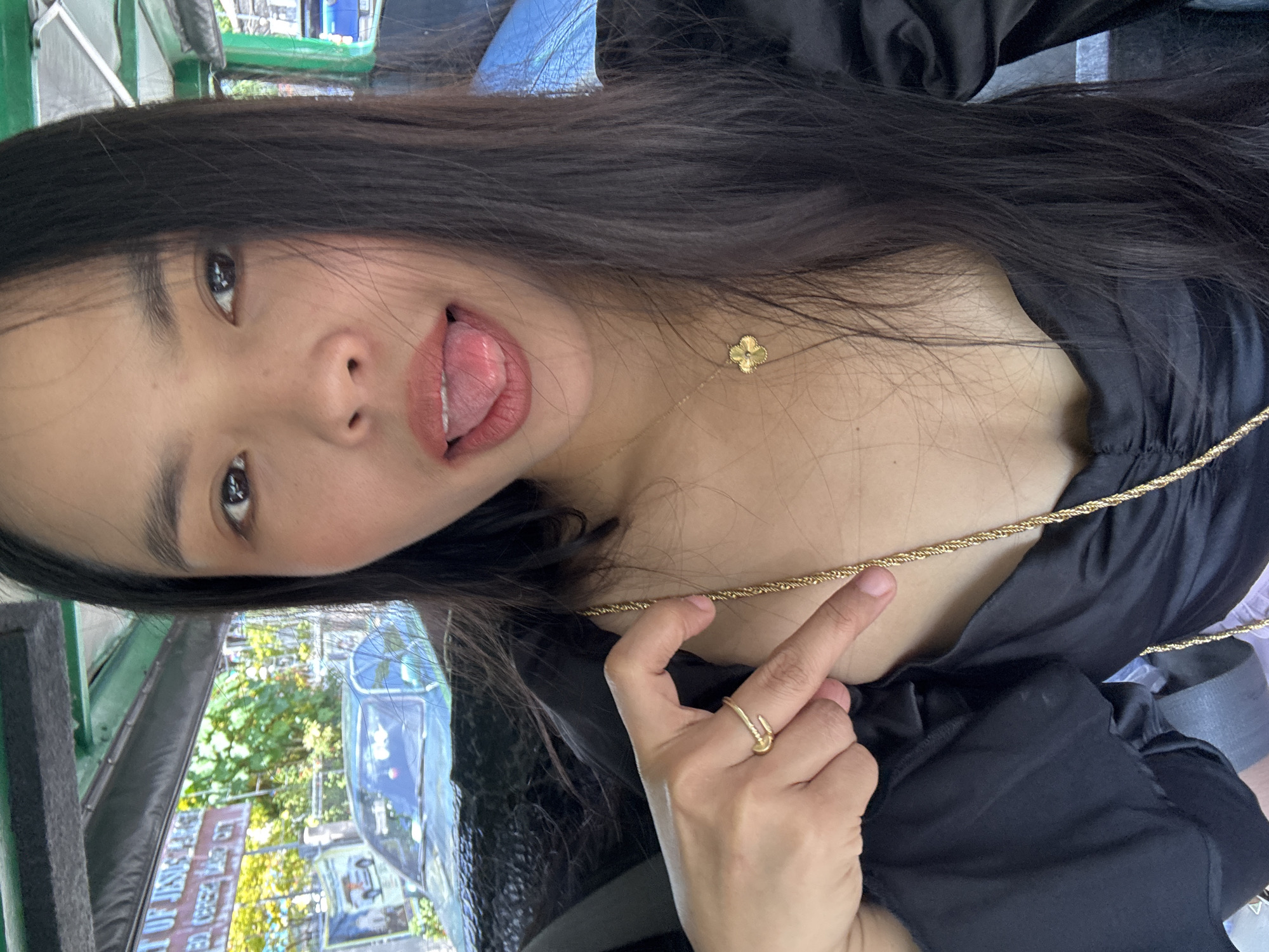 A close-up, sideways portrait captures a young woman, likely of East Asian descent, within what appears to be an urban setting. She has long, dark hair and wears a black top, possibly with ruffled or off-the-shoulder detailing. Her expression is playful, with her tongue slightly extended from her mouth, and her dark eyes are directed towards the viewer.
She is accessorized with two gold necklaces: a delicate chain featuring a small four-leaf clover pendant and a thicker, rope-style gold chain. A distinctive gold ring, designed to resemble a nail, is worn on her right ring finger. Her right hand is visible near her chest, gently holding the thicker necklace.
The background shows distorted reflections on a surface, possibly a vehicle window, suggesting an outdoor, daytime scene with bright natural light. These reflections include urban elements such as buildings, lush green trees, and other vehicles, consistent with a bustling city environment. Faint, inverted text, possibly "JUSTICE," can be discerned within these reflections, but it is heavily distorted. The context of Davao City, Philippines, aligns with the visual cues of an active urban landscape and potential local transport elements in the background. The woman's pose and playful expression contribute to a lighthearted mood.