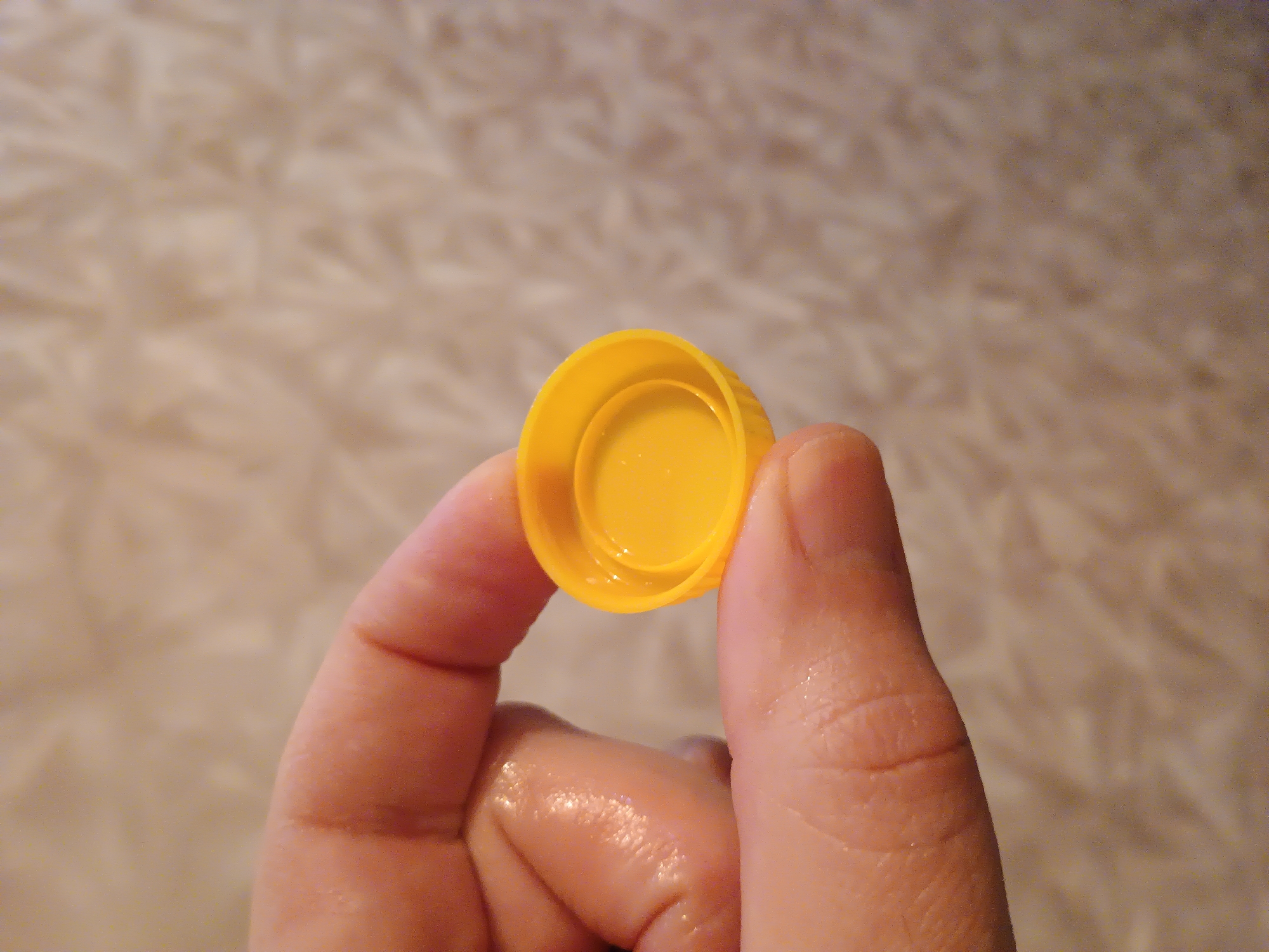 A close-up shot shows a person's hand holding a yellow plastic bottle cap. The cap has a ribbed exterior and a recessed interior, with a smaller circular indentation within that. The hand, with visible fingerprints and slight sheen from moisture, grips the cap between the thumb and index finger. The background consists of a softly textured, light-colored surface, possibly fabric or carpet. The scene is devoid of any identifying markers of Zaporizhzhia, Ukraine.